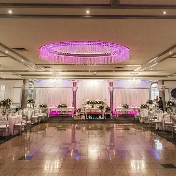 The image shows an elegantly decorated indoor wedding venue provided by Buffet Marina, categorized under Wedding Venues for Mariage Elegant in Canada. The spacious hall features a polished marble floor and is illuminated by modern recessed lighting, accented with a stunning circular chandelier emitting a pink hue. The room is arranged with round tables set for guests, each adorned with white tablecloths and elegant floral centerpieces. Silver chiavari chairs surround the tables, complementing the sophisticated ambiance. At the far end of the hall, a decorative backdrop highlights a long, flower-adorned head table, creating a focal point for the wedding celebration.