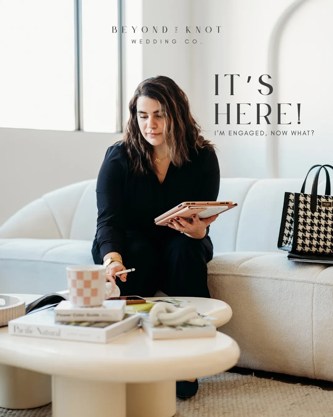 In the image, a woman is seated on a light-colored sofa in a modern, stylish room. She is holding a tablet in one hand, appearing focused and engaged in her work. On the table in front of her are open books and a checkered mug, suggesting a planning session or meeting. The room has large windows allowing natural light to flood in, highlighting the chic, elegant decor. A houndstooth-patterned bag is placed on the couch beside her. Above, the text reads, 'It's Here! I'm Engaged, Now What?' This image represents the services of Beyond the Knot, categorized under Wedding Planners for Mariage Elegant in Canada, emphasizing professional planning and consultation for newly engaged couples.
