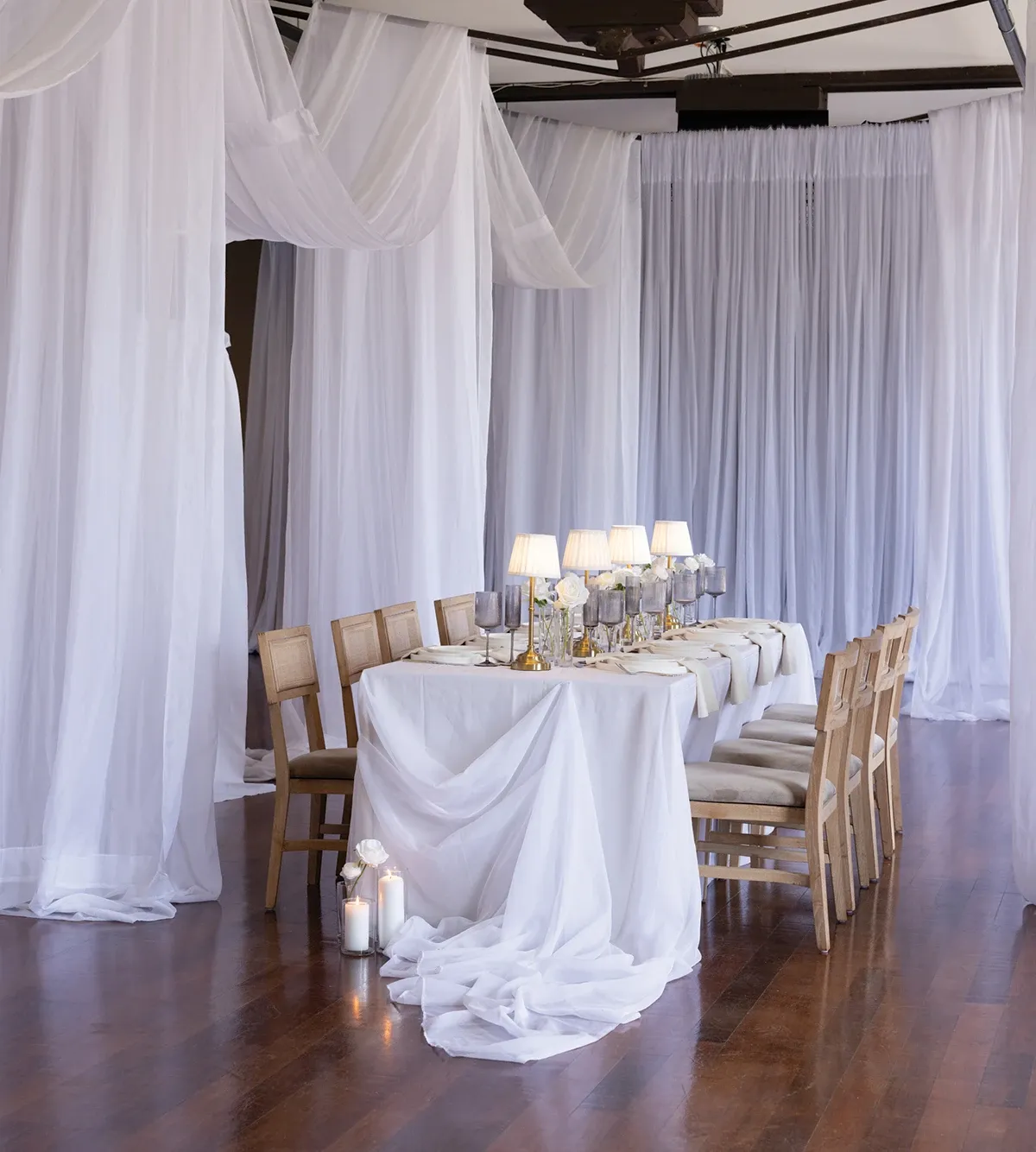 The image showcases an elegantly arranged wedding reception table, curated by the wedding planners at Beyond the Knot, categorized as Wedding Planners on Mariage Elegant, Canada. The table is draped in soft white linens, complemented by a series of illuminated table lamps that cast a warm, ambient glow. Wooden chairs are neatly aligned along both sides of the table. In the background, sheer white curtains add a touch of sophistication and intimacy to the setting. The polished wooden floor enhances the elegance of the overall arrangement, ideal for an upscale, romantic wedding reception.