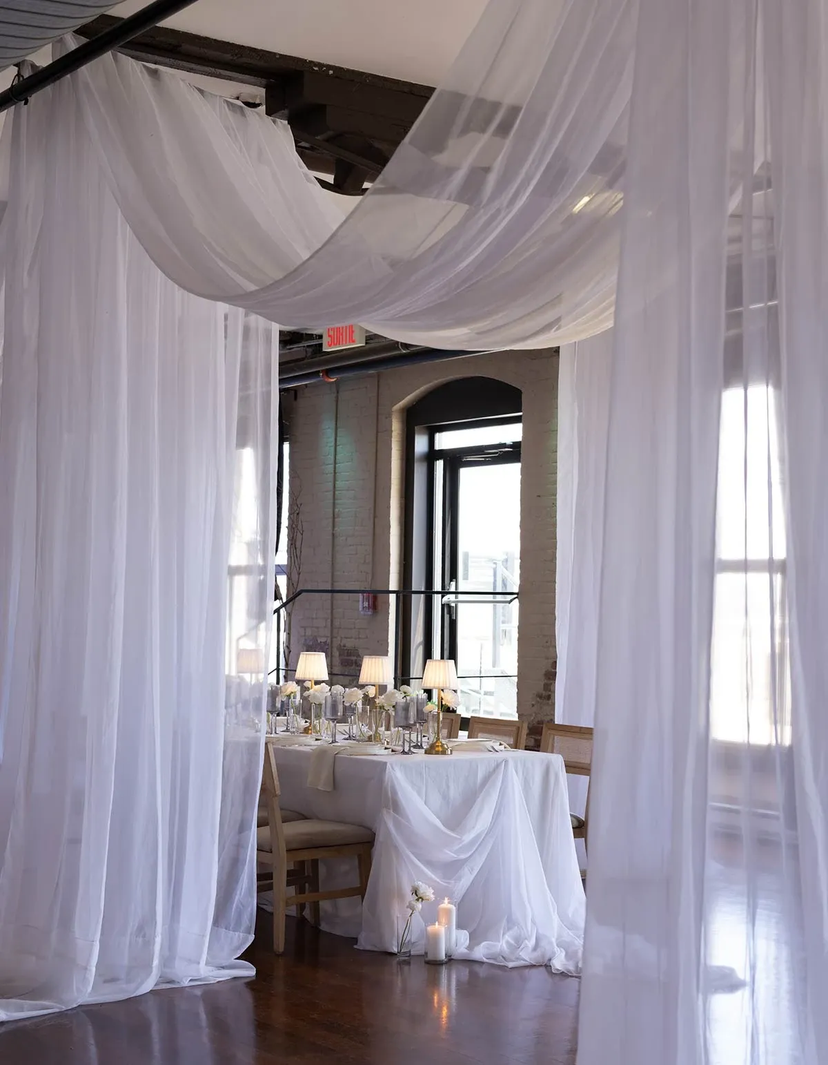 The image showcases an elegantly decorated wedding reception scene by Beyond the Knot, a wedding planner in the Wedding Planners category. The setting features softly draped sheer white curtains creating a romantic, intimate atmosphere. A beautifully arranged table is at the center, adorned with white tablecloths and various glass candle holders, contributing to a warm, inviting glow. Stylish chairs are arranged around the table, and dainty lamps provide additional lighting, enhancing the sophisticated ambiance ideal for upscale weddings.