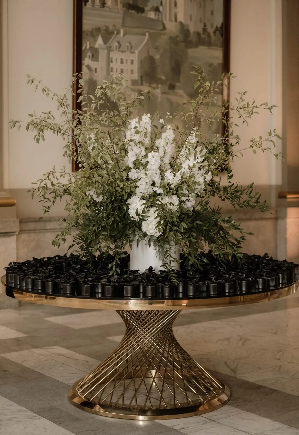 In a sophisticated interior setting, an elegant gold wireframe table is prominently placed, showcasing Beyond the Knot's expertise in the Wedding Planners category. The table is adorned with an ornate floral arrangement featuring lush green leaves and tall white blossoms, subtly highlighting the vendor's attention to detail and design. Surrounding the floral centerpiece, a meticulously arranged circle of small black boxes adds a touch of modern luxury, perfect for a refined wedding celebration. The backdrop features a grand painting, enhancing the sophisticated ambiance of the scene.