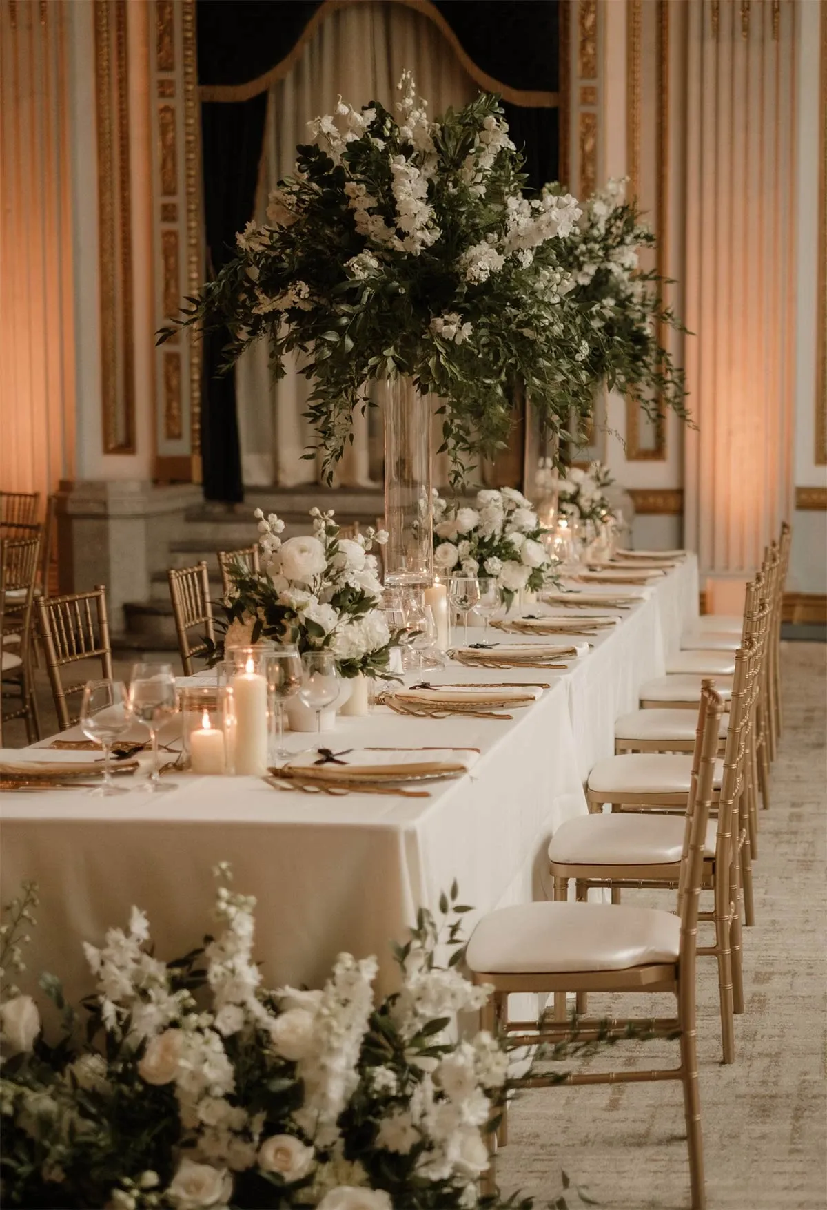 The image features an elegantly decorated wedding reception table, showcasing the sophisticated planning skills of Beyond the Knot, a wedding planner in Canada. The long, rectangular table is adorned with a pristine white tablecloth, and each place setting is accentuated with a gold charger plate and polished silverware. At the center of the table, tall glass vases hold abundant arrangements of white and green florals, adding a lush and fresh ambiance. Soft candlelight enhances the romantic atmosphere, with several lit candles placed strategically along the table. Gold chiavari chairs provide elegant seating, complementing the luxurious setting. The backdrop features opulent drapery and intricate architectural details, creating a grand and inviting setting perfect for a memorable wedding celebration.