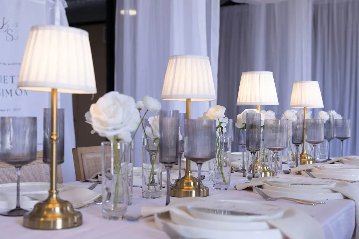 The image showcases an elegant wedding table setting arranged by Beyond the Knot, a vendor in the Wedding Planners category. The table features a series of stylish, gold-colored lamps with white shades, casting a warm glow over the arrangement. Each setting includes delicate white napkins placed on white plates, complemented by tall, grey-tinted wine glasses and crystal-clear water glasses. White roses in slender glass vases are interspersed between the lamps, adding a touch of sophistication. The background displays soft, white draped fabric, creating an overall elegant and serene atmosphere perfect for a wedding celebration.