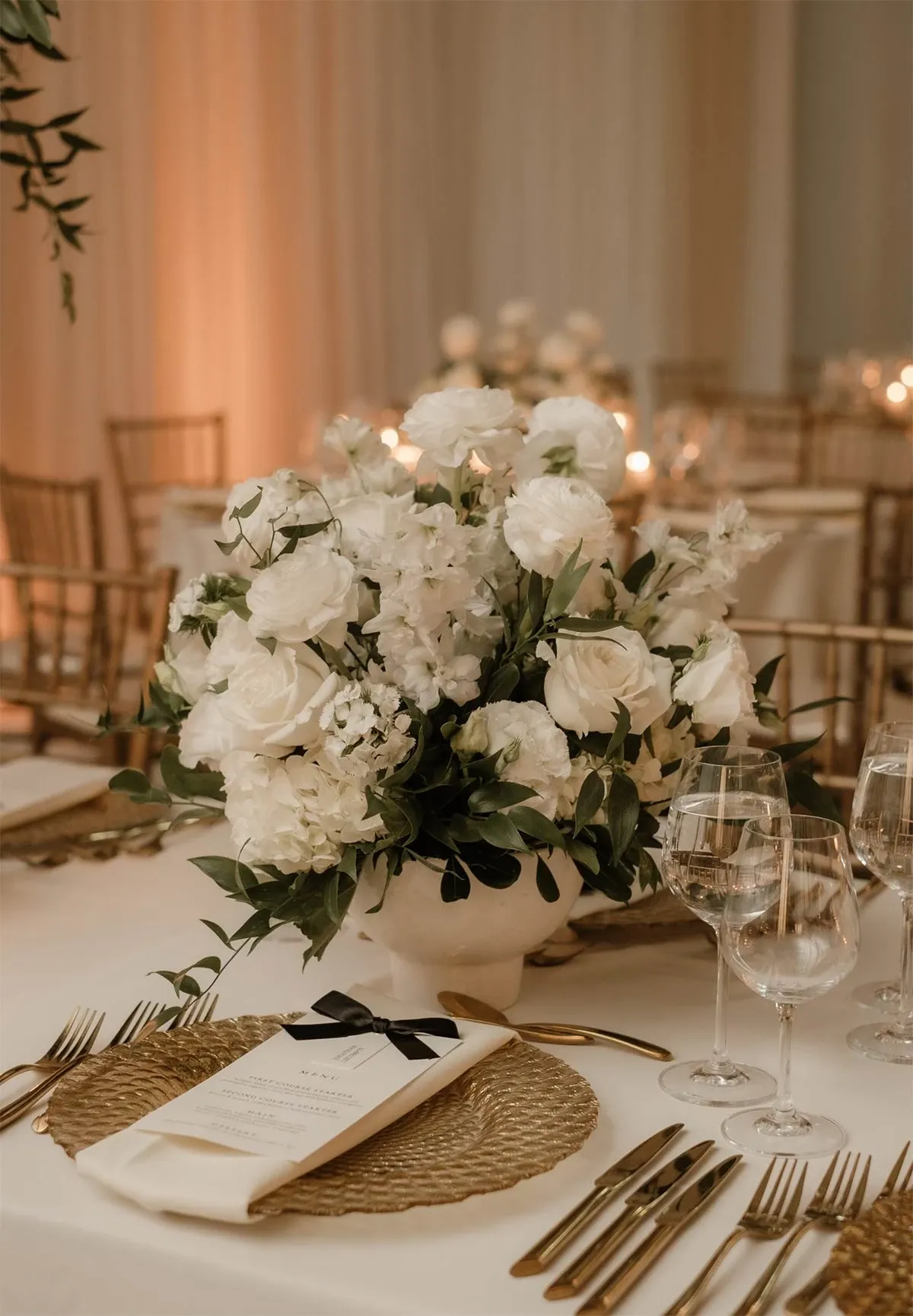 The image showcases a beautifully set wedding reception table by Beyond the Knot, a reputable wedding planner listed under the Wedding Planners category on Mariage Elegant, Canada. The table features an elegant centerpiece of white roses and peonies, complemented by lush greenery in a classic white vase. Surrounding the arrangement are gold-rimmed wine glasses and a neatly folded napkin with a black ribbon, resting on a round woven placemat. The soft lighting and wooden chairs in the background create a warm, inviting atmosphere, ideal for a sophisticated wedding celebration.