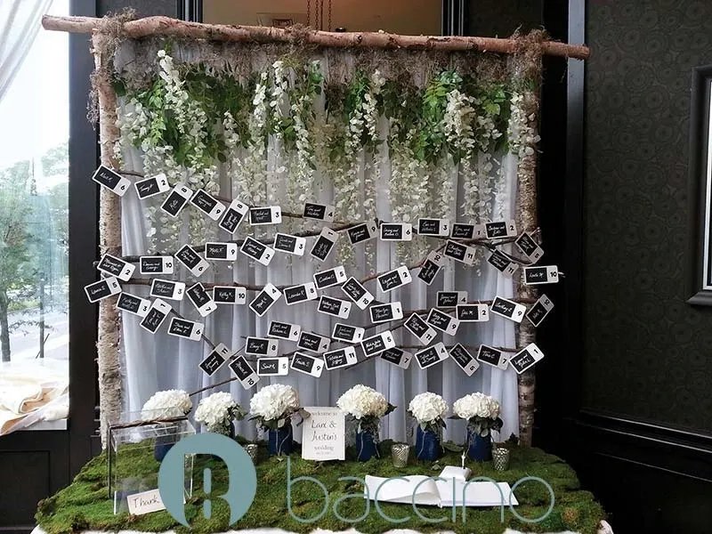 This image showcases a beautifully arranged wedding setup by Baccino Events, falling under the Wedding Planners category. It features an elegant display of cascading white and green floral arrangements hanging from a rustic wooden frame. In front of the floral backdrop, numerous place cards are artistically pinned on strings, possibly for guest seating arrangements. Below, several white hydrangea bouquets are placed in blue glass vases on a table covered with a lush green moss-like surface. This stylish and organic arrangement captures the sophistication and attention to detail that Baccino Events brings to wedding planning in Canada.