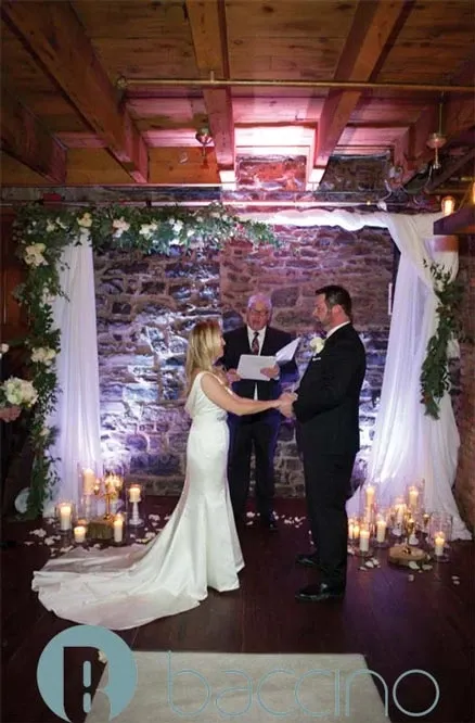 A bride and groom stand facing each other under a rustic wedding arch adorned with greenery and white drapery in an intimate, warmly lit setting. The stone wall backdrop adds an elegant touch, complemented by numerous lit candles arranged around the couple, creating a romantic atmosphere. An officiant stands between them, holding a booklet, ready to conduct the ceremony. This scene showcases the meticulous planning and decor provided by Baccino Events, captured under their wedding planners category on Mariage Elegant, Canada.