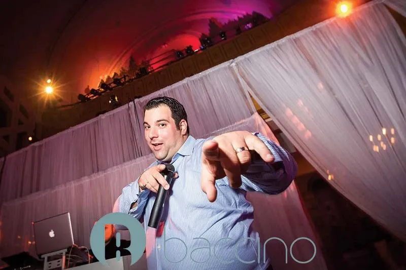 A wedding planner from Baccino Events, featured in a dynamic setting, stands with a microphone in hand, engaging the crowd. The person is dressed in a light blue shirt and is in front of an elegant backdrop consisting of sheer white drapery cascading down. The venue has soft, ambient lighting with a warm glow, typical for a wedding celebration. This image represents Baccino Events in the wedding planners category for Mariage Elegant, Canada, showcasing their lively and interactive approach to event coordination.