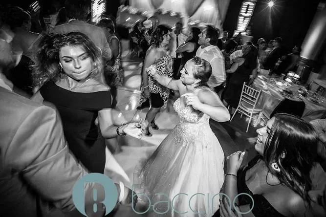 This black and white image captures a lively dance floor at a wedding reception, managed by Baccino Events, classified under Wedding Planners. Guests are energetically dancing, showcasing joy and celebration. The bride, wearing an elegant gown, is at the center, smiling and enjoying the moment. Surrounding her are well-dressed guests, both men and women, participating in the festivities. The ambiance is enhanced by dynamic lighting in the background and elegantly set tables with white chairs, suggesting a stylish and well-organized event by Baccino Events.