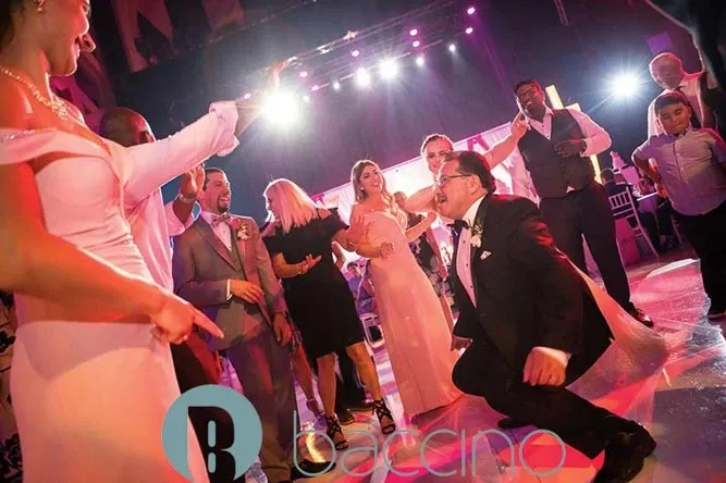 A lively wedding reception scene featuring Baccino Events, a renowned wedding planner in Canada, showing guests dancing energetically on a well-lit dance floor. The photograph captures elegantly dressed attendees, including a man in a suit joyfully squatting down in front of a woman in a pink dress. The background showcases vibrant lighting that enhances the festive atmosphere. This image reflects the expertise of Baccino Events in creating memorable wedding experiences, as highlighted on Mariage Elegant's wedding planning and vendor listing.