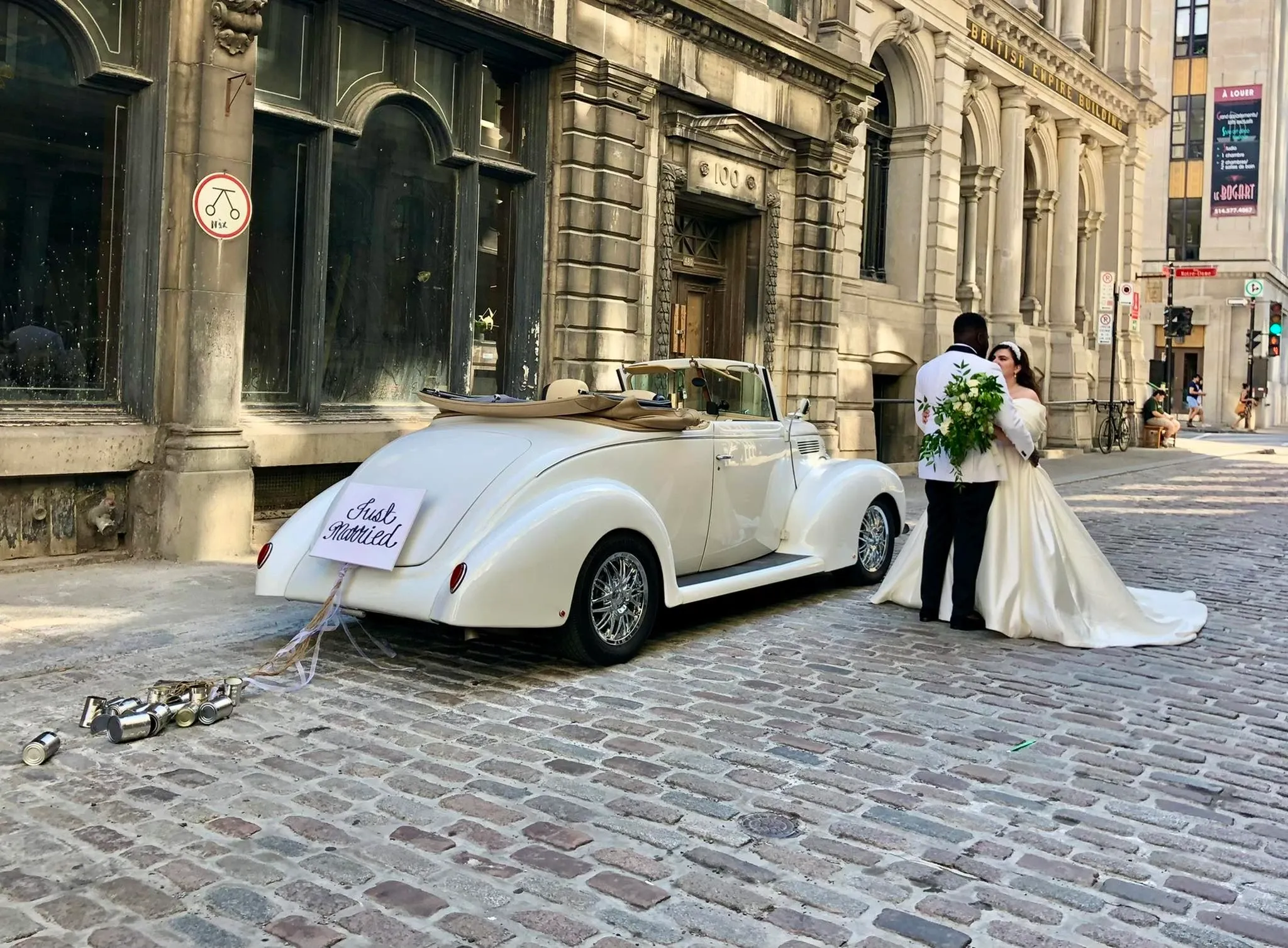 The image features a classic white convertible limousine by Antique Limousine, categorized under Wedding Limousine, parked on a cobblestone street. The elegant vehicle is adorned with a 'Just Married' sign at the back, along with strings of decorative tin cans trailing behind. A newlywed couple stands nearby, embracing lovingly; the bride wears a flowing white gown and holds a vibrant bouquet, while the groom is dressed in a formal suit. The historic buildings lining the street add an old-world charm, enhancing the romantic and sophisticated atmosphere suitable for a wedding celebration in Canada.