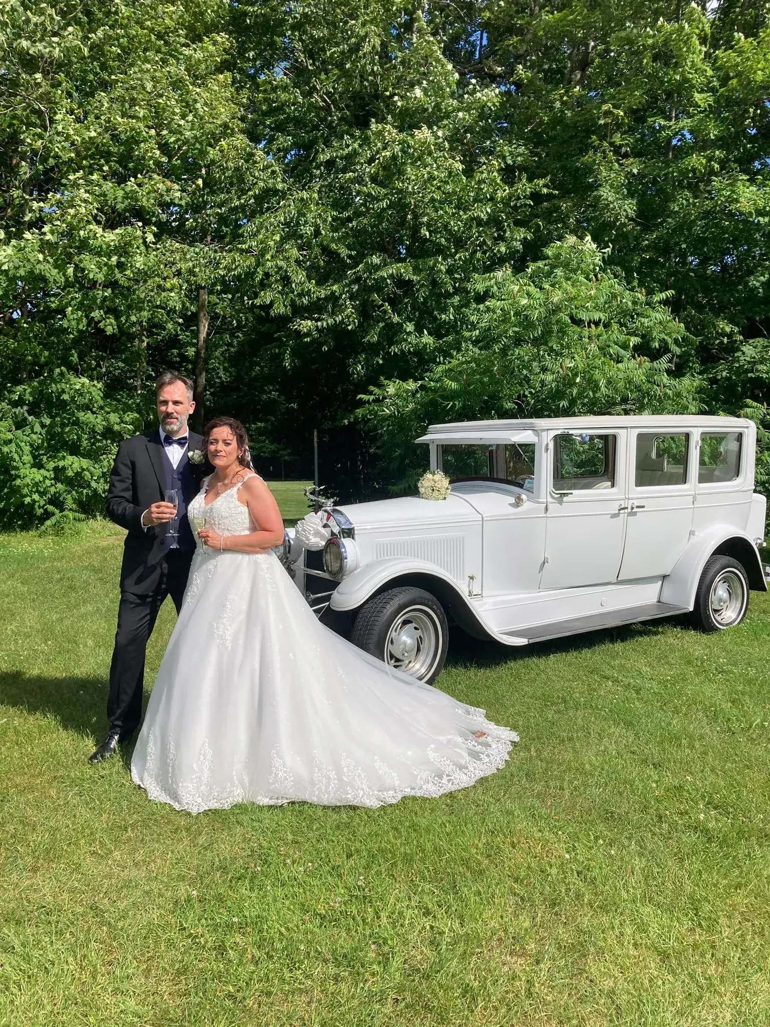 In this image on the Mariage Elegant website, a bride and groom stand beside an elegant, vintage white car, provided by the vendor Antique Limousine, under the category of Wedding Limousine. The bride is wearing a beautiful white gown with intricate lace detailing, while the groom is dressed in a classic black tuxedo with a bow tie. The couple appears joyful, holding champagne glasses, set against a lush green background, enhancing the romantic and celebratory atmosphere of their wedding day. The vintage car, adorned minimally with white flowers, adds a touch of timeless elegance to the scene, making it perfect for their special occasion.