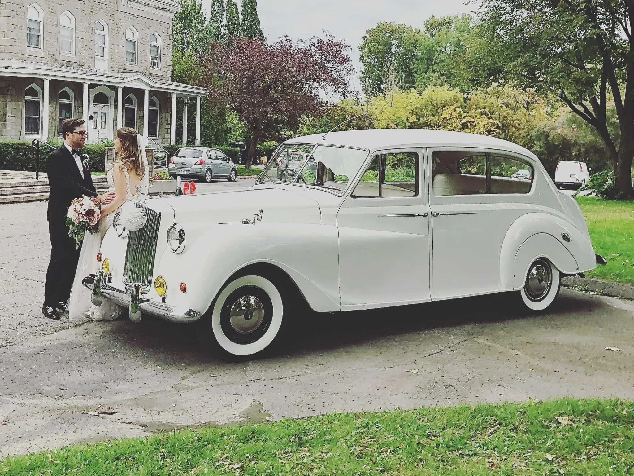 In the image, a classic white vintage limousine, provided by Antique Limousine, is parked gracefully in front of a historic stone mansion, surrounded by lush greenery. It fits perfectly in the wedding limousine category, capturing the timeless elegance often sought in wedding transport. In the foreground, a newlywed couple stands beside the car — the groom is wearing a traditional black tuxedo while the bride holds a bouquet and wears a flowing white gown. The scene conveys an air of sophistication and romance, ideal for couples seeking a memorable wedding day experience.