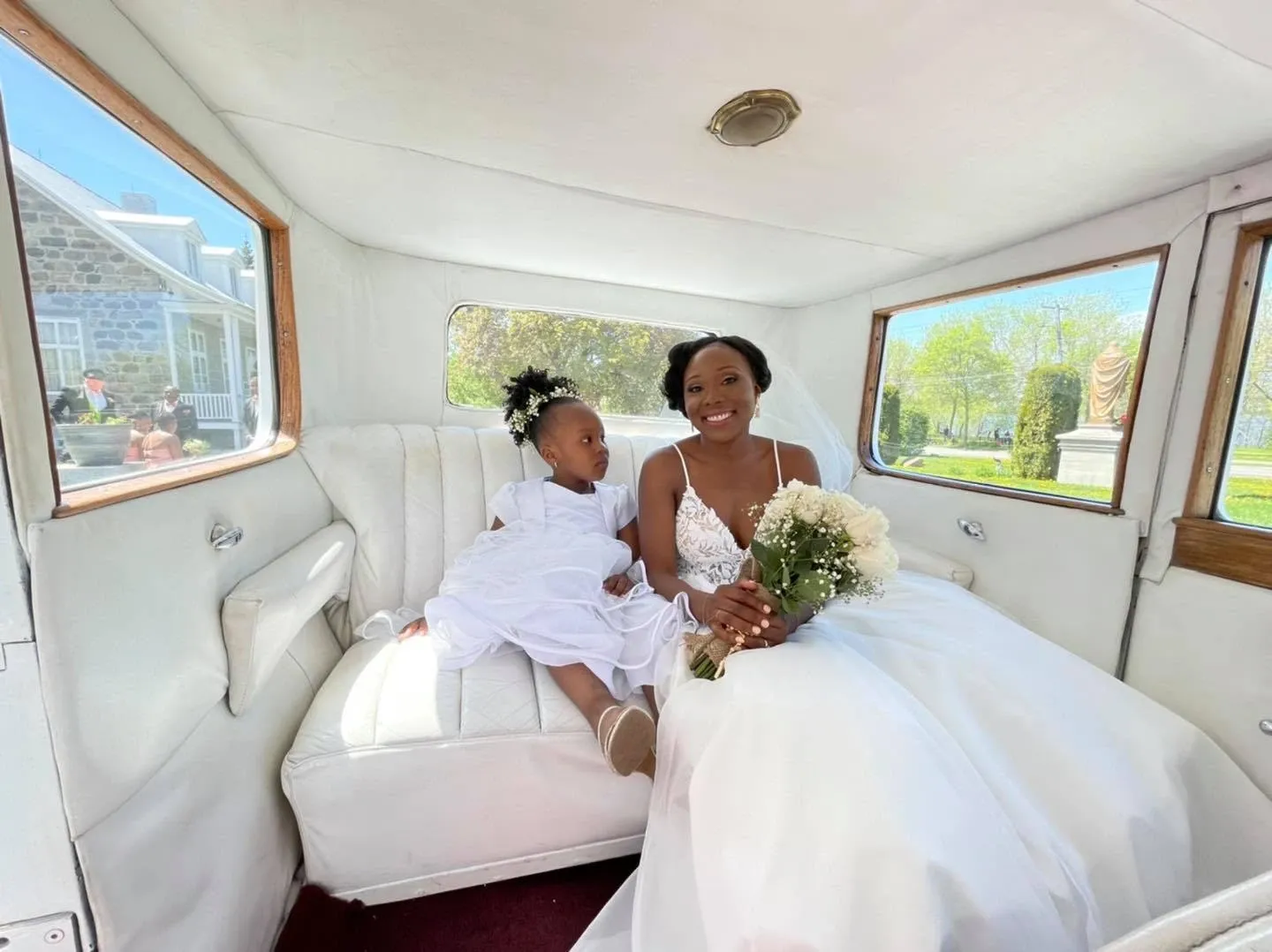A joyful bride and a young girl, both in elegant white dresses, are seated inside a beautifully restored vintage limousine provided by Antique Limousine for a wedding. The bride is holding a bouquet of white flowers and smiling brightly, capturing the essence of her special day. The interior of the limousine features plush white seating, exuding a classic and luxurious ambiance. Through the windows, a sunny day and a glimpse of a stone building surrounded by greenery can be seen, adding to the charm and elegance of the wedding setting. The image highlights Antique Limousine’s dedication to providing a sophisticated and memorable transportation experience for weddings.