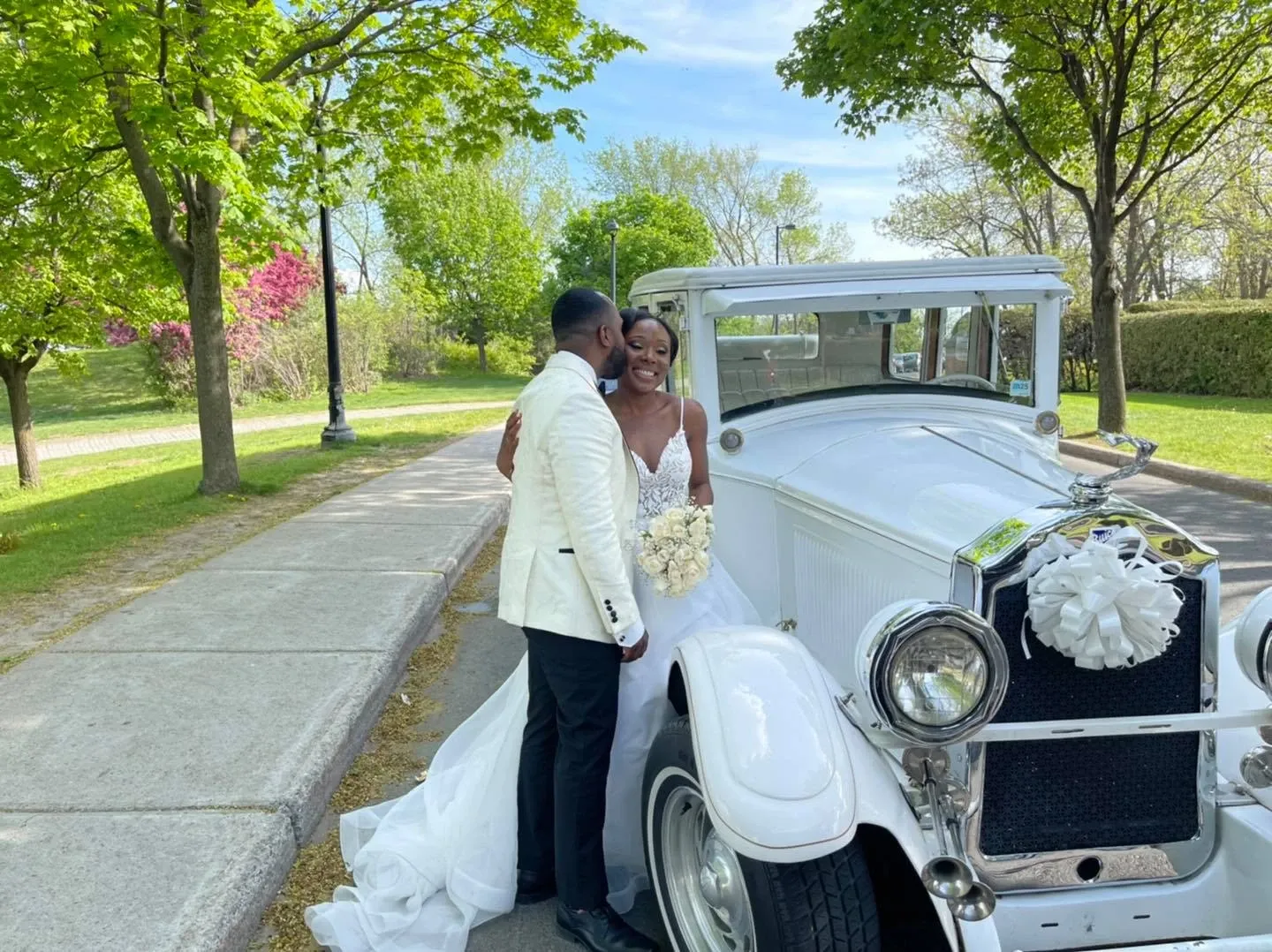 A happy couple stands beside a classic white limousine from Antique Limousine, featured in the wedding limousine category on Mariage Elegant, Canada. The bride, holding a bouquet of white roses, is dressed in a stunning white gown with a long train, while the groom wears an elegant white tuxedo with a black bow tie. They are embracing in a picturesque park setting, surrounded by lush green trees and a clear blue sky, capturing the essence of a perfect wedding day. The vintage limousine, adorned with a white ribbon and flower decoration on the front, adds a touch of timeless elegance to the scene.