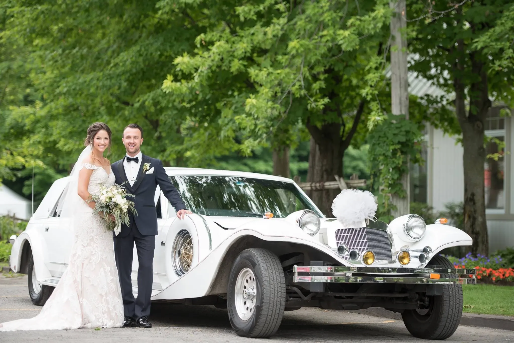 A bride and groom stand beside an elegant, vintage-style white limousine provided by Antique Limousine, a vendor in the Wedding Limousine category. The groom is dressed in a black tuxedo, and the bride wears a beautiful white gown, holding a bouquet of flowers. The limousine features striking chrome details and a distinctive, classic design, parked on a picturesque lane surrounded by lush green trees, enhancing the idyllic wedding setting. This image captures a moment of sophistication and romance, perfect for a memorable wedding day in Canada.