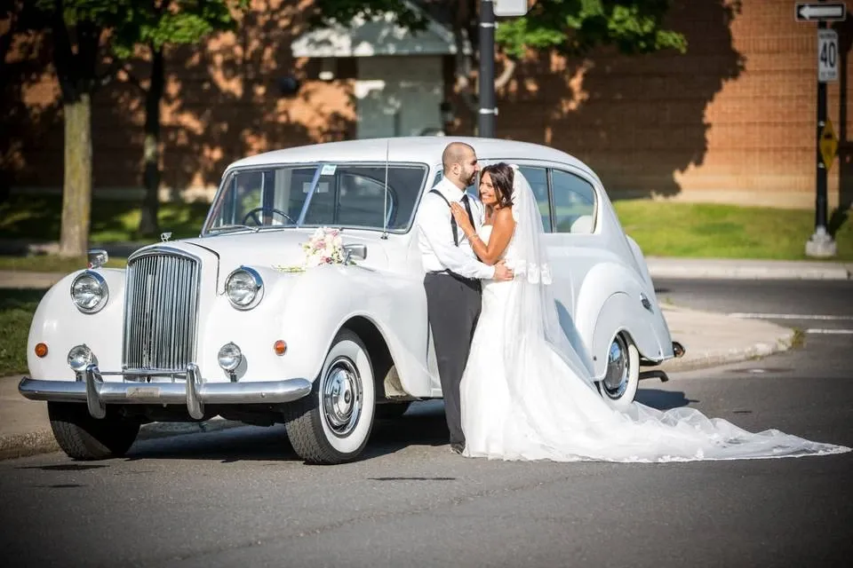 A bride and groom stand happily in front of a classic white vintage limousine provided by Antique Limousine, ideal for wedding transportation. The couple is elegantly dressed, with the bride in a flowing white gown and veil, and the groom in a formal suit with a vest. The limousine, categorized under wedding limousine, features polished chrome details and is adorned with a small floral arrangement on the front. The sunny day, along with lush green trees and a brick building in the background, adds to the joyous occasion. This scene is perfect for highlighting the charm and sophistication that Antique Limousine can bring to any wedding day in Canada.