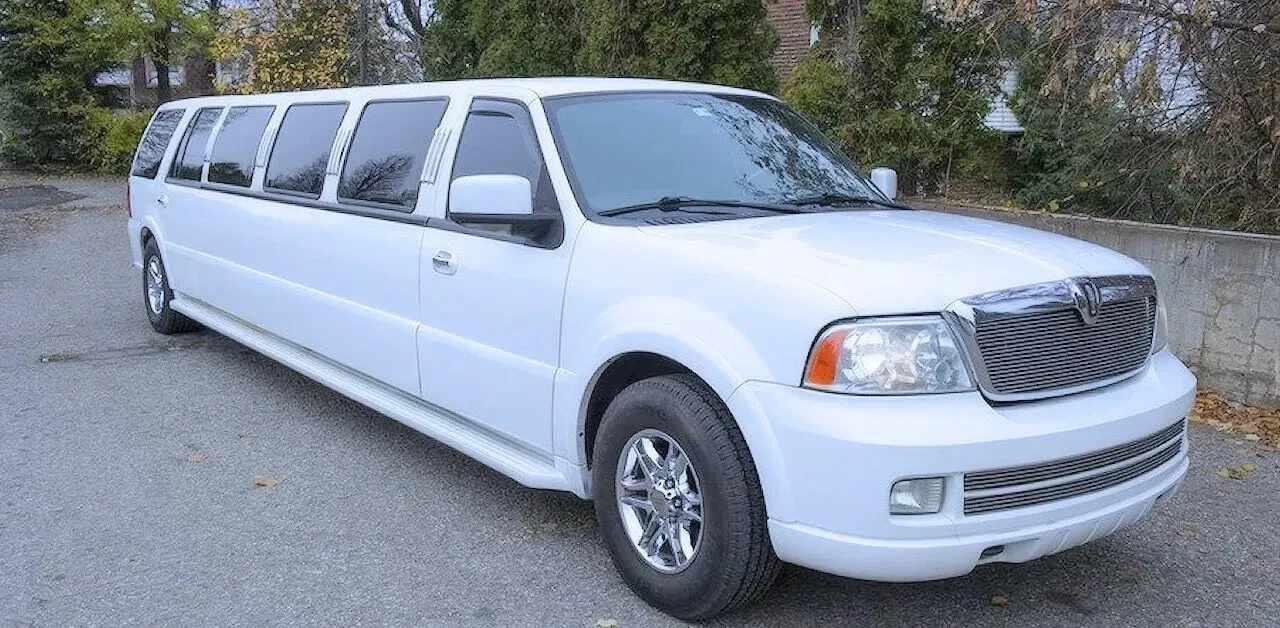 The image shows a luxurious, white stretch limousine provided by Antique Limousine, categorized under Wedding Limousine services. The vehicle is parked on a paved surface with trees in the background, showcasing its sleek design and polished exterior. The limousine features shiny chrome wheels and tinted windows, adding to its elegant appearance, which is ideal for wedding celebrations. This setting suggests a perfect mode of transportation for a bridal party or newlyweds looking for classic and stylish wedding day travel in Canada.