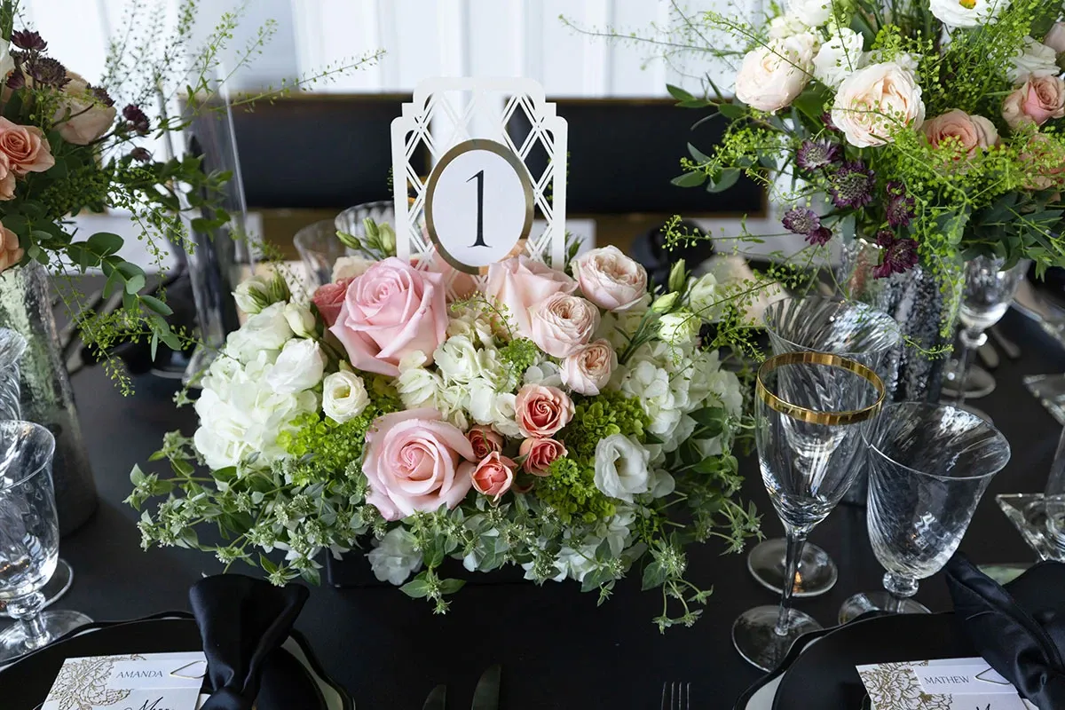 The image showcases a beautifully arranged centerpiece by Ombelle Fleuriste, categorized under Wedding Florists, for a wedding setting. The floral arrangement features an elegant display of pink and white roses, accented with green foliage and delicate white blossoms, creating a romantic and sophisticated ambiance. Crystal glassware with gold rims and neatly folded black napkins complement the floral design on a dark tablecloth, while a table number '1' card is prominently placed within the arrangement, signifying an organized seating arrangement for a wedding reception.