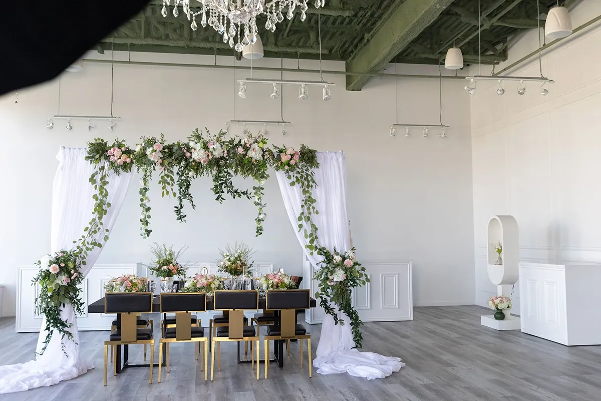 The image showcases a beautifully arranged wedding setup by Ombelle Fleuriste, categorized under Wedding Florists, at Mariage Elegant in Canada. The center of attention is a stunning floral arch adorned with lush greenery and pastel flowers, elegantly draped with white fabric. Below the arch is a sophisticated dining area featuring a long table with black and gold chairs, each place setting complemented with floral centerpieces that echo the arch's design. The decor creates a harmonious and elegant atmosphere, perfect for a wedding celebration.