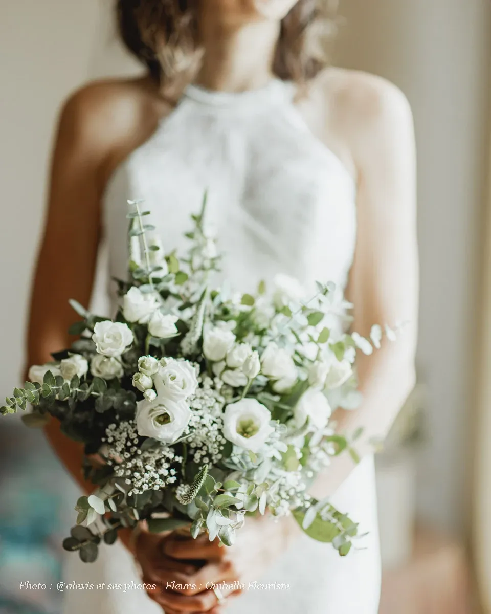 A bride in a sleeveless, high-neck white wedding gown gently holds an elegant bouquet designed by Ombelle Fleuriste, categorized under wedding florists. The bouquet is composed of delicate white roses, baby's breath, and lush eucalyptus leaves, creating a classic and sophisticated floral arrangement perfect for weddings. The soft-focus background adds a dreamy ambiance to the scene.