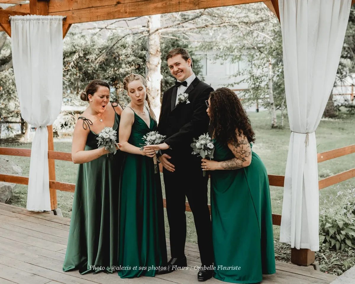 The image features a joyful wedding party on a wooden deck surrounded by lush greenery. Four individuals, three bridesmaids in elegant green dresses and a groom in a classic black suit, are posed playfully. Each bridesmaid holds a small bouquet of white flowers, showcasing the floral arrangements provided by Ombelle Fleuriste, specializing in wedding floristry. The setting is bright and natural, complementing the wedding theme and enhancing the occasion's elegance. Ombelle Fleuriste's floral design adds a touch of beauty and coordination to the celebration.