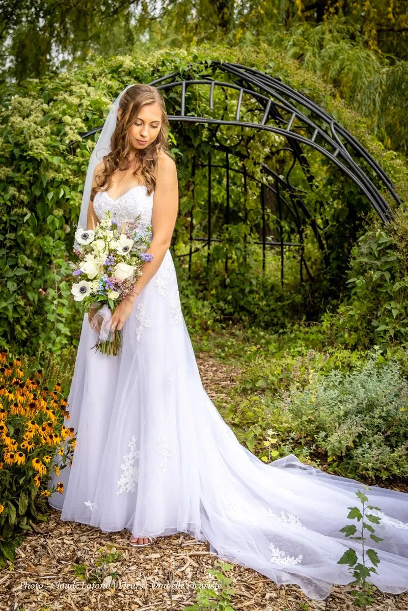 The image features a bride standing in a lush garden setting, showcasing Ombelle Fleuriste's expertise as a wedding florist. She is elegantly dressed in a white lace wedding gown, holding a beautifully arranged bouquet. The bouquet consists of white and purple flowers, complemented by greenery, offering a romantic and sophisticated touch. In the background, there is a charming wrought iron arch adorned with trailing plants and surrounded by vibrant greenery, creating a picturesque wedding scene. This setting highlights Ombelle Fleuriste’s skill in creating stunning wedding floral arrangements.