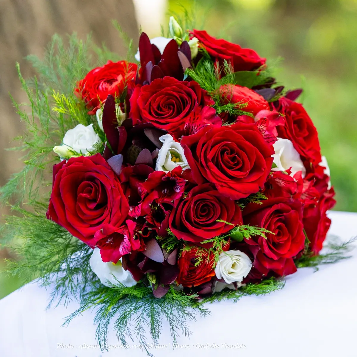 This image showcases a vibrant bridal bouquet crafted by Ombelle Fleuriste, a vendor in the wedding florists category. The bouquet is artfully arranged with an array of lush red roses, delicate white flowers, and deep maroon blooms, creating a striking contrast. Accented with greenery and fern-like leaves, it rests elegantly on a white surface, set against a blurred natural background. This floral arrangement epitomizes the sophistication and beauty essential for a wedding, reflecting the expertise of Ombelle Fleuriste in creating memorable floral displays for special occasions.