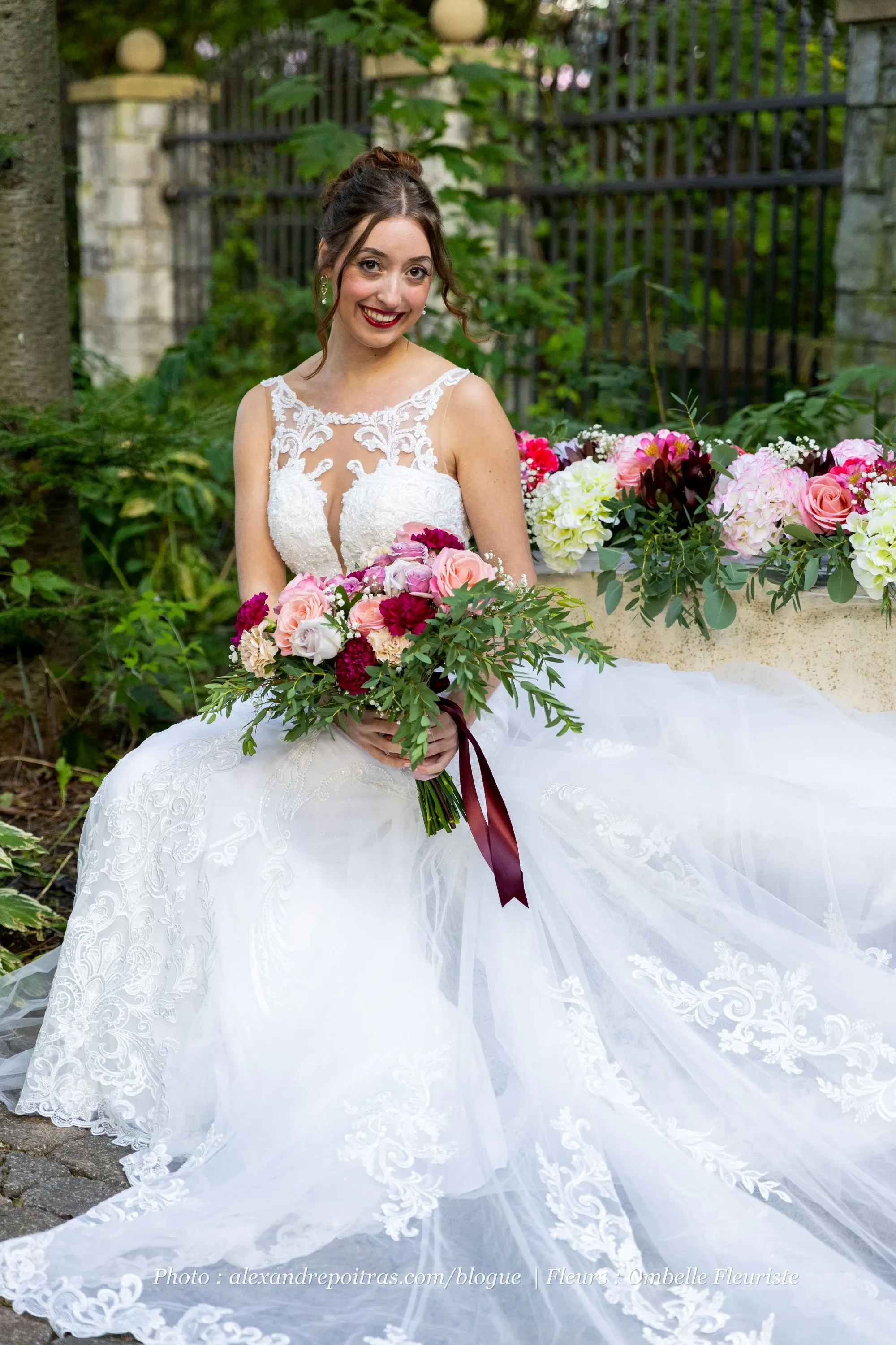 In this image, a bride is seated outdoors, wearing an elegant white lace wedding gown with intricate embroidery. She is holding a vibrant bouquet composed of pink and red flowers, accented with lush greenery. The setting has a natural feel, with greenery in the background and a stone bench adorned with additional floral arrangements, featuring roses and hydrangeas in shades of pink, white, and red. This beautiful floral display is crafted by Ombelle Fleuriste, specialists in wedding florists for Mariage Elegant in Canada. The scene exudes romance and elegance, perfectly capturing the essence of a wedding celebration.