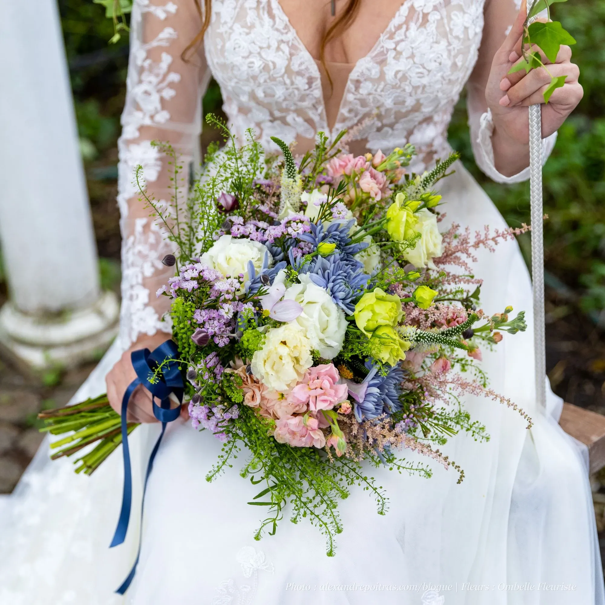 In this image, a bride in an elegant lace wedding gown holds a vibrant floral bouquet created by Ombelle Fleuriste, a wedding florist listed on Mariage Elegant, Canada. The bouquet features a stunning mix of white, pink, purple, and blue flowers, beautifully complemented with lush green foliage. The intricate lace details of the bride's dress and the colorful arrangement add a touch of elegance and romance, ideal for any wedding setting.