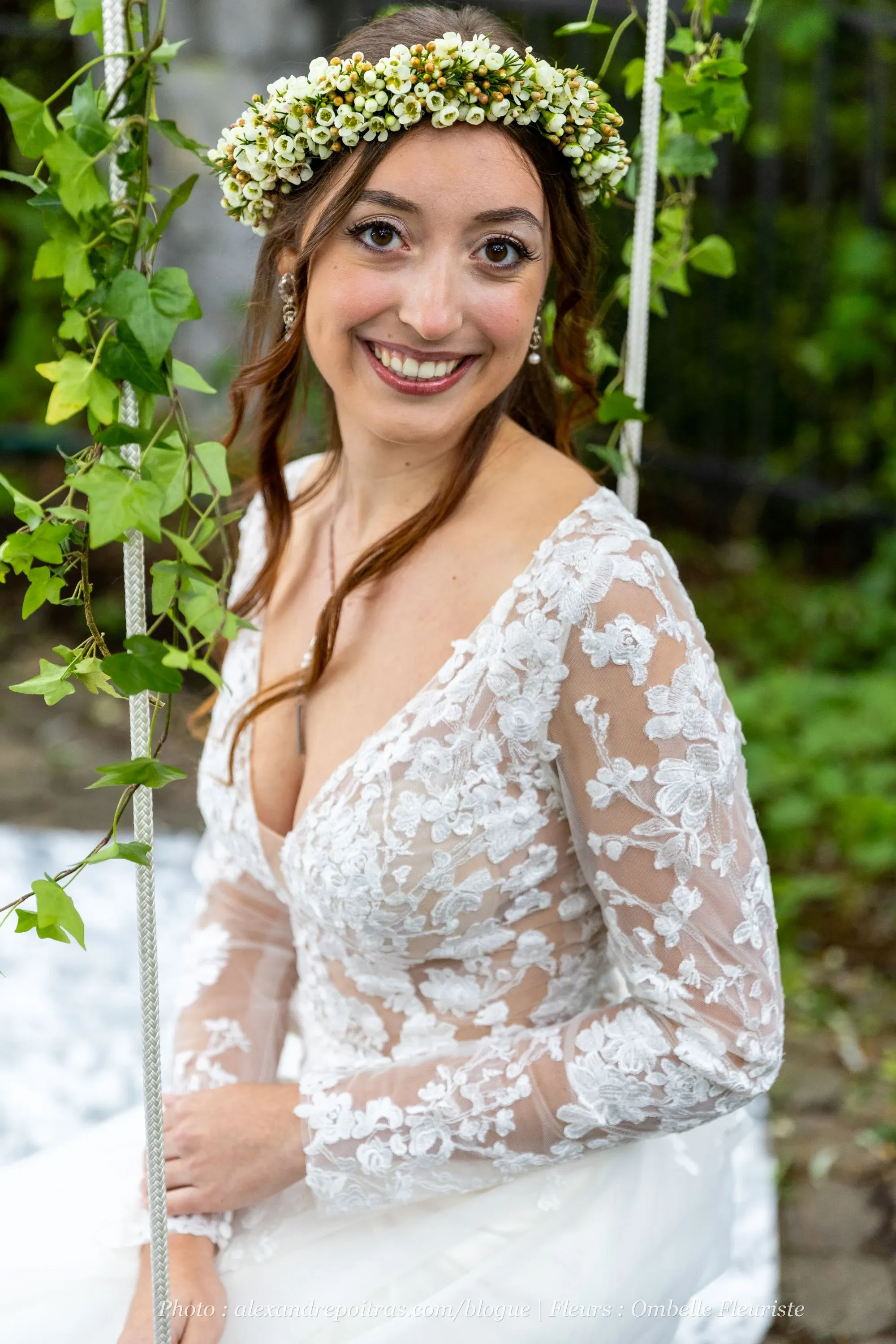 A bride is seated on a wooden swing adorned with lush green ivy, wearing an elegant, long-sleeved lace wedding dress featuring intricate floral patterns. She smiles softly, gazing at the camera. Her long hair is styled in gentle waves, and she wears a delicate floral crown made of small white flowers. The background hints at an outdoor setting, enhancing the natural and romantic ambiance. This image is associated with Ombelle Fleuriste, a wedding florist listed on Mariage Elegant in Canada.