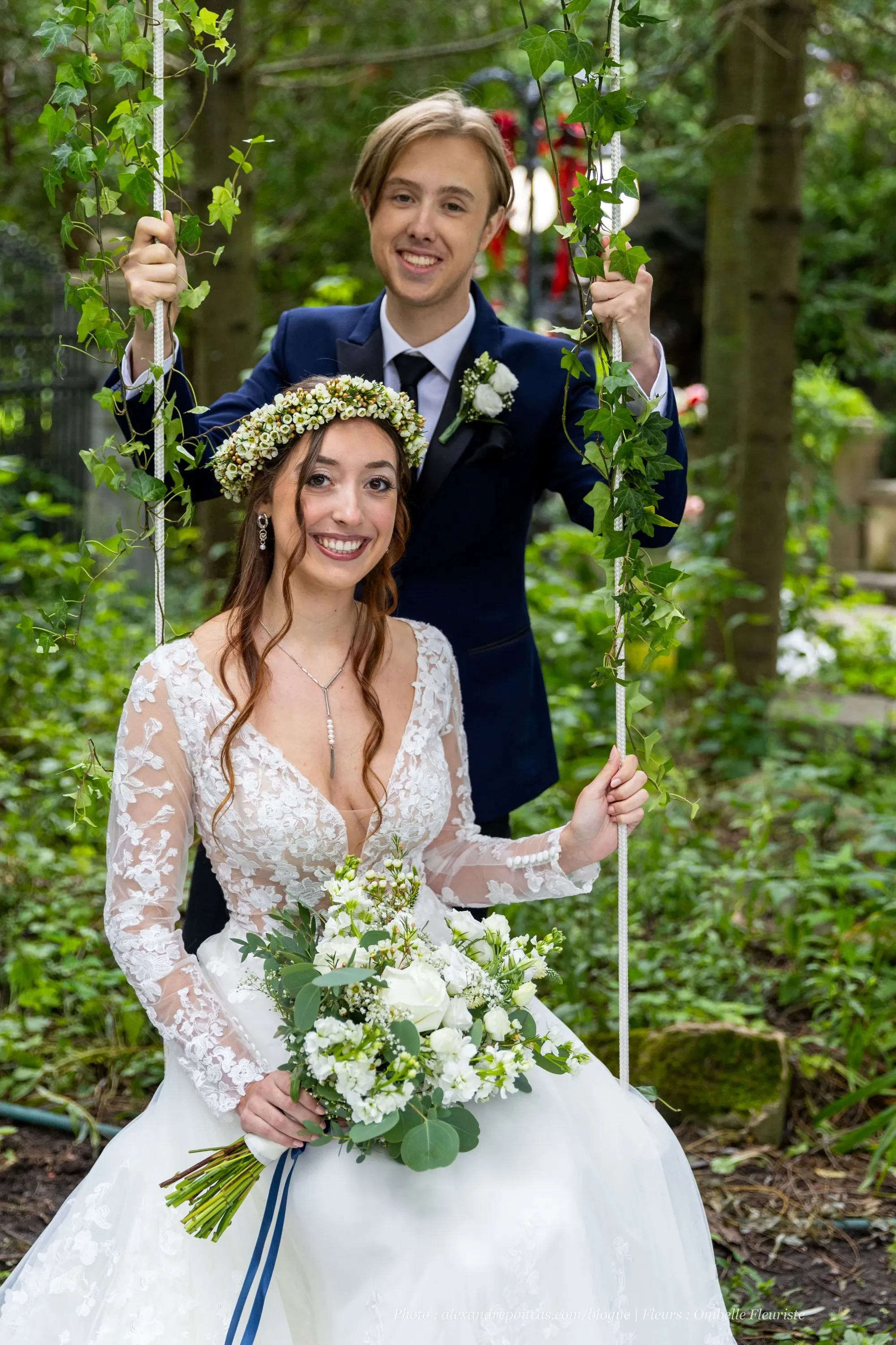 In a lush, green outdoor setting, a bride and groom are joyfully posing on a swing adorned with greenery. The bride, dressed in an elegant white lace wedding gown, holds a bouquet of white flowers and greenery, created by Ombelle Fleuriste. Her hair is decorated with a delicate floral crown, adding a natural, romantic touch to her look. The groom stands behind her, dressed in a classic navy suit with a boutonniere matching the bride's bouquet. This image, depicting the artistry of a wedding florist, showcases the intricate floral design and styling by Ombelle Fleuriste, highlighting their talent in creating beautiful floral arrangements for weddings.