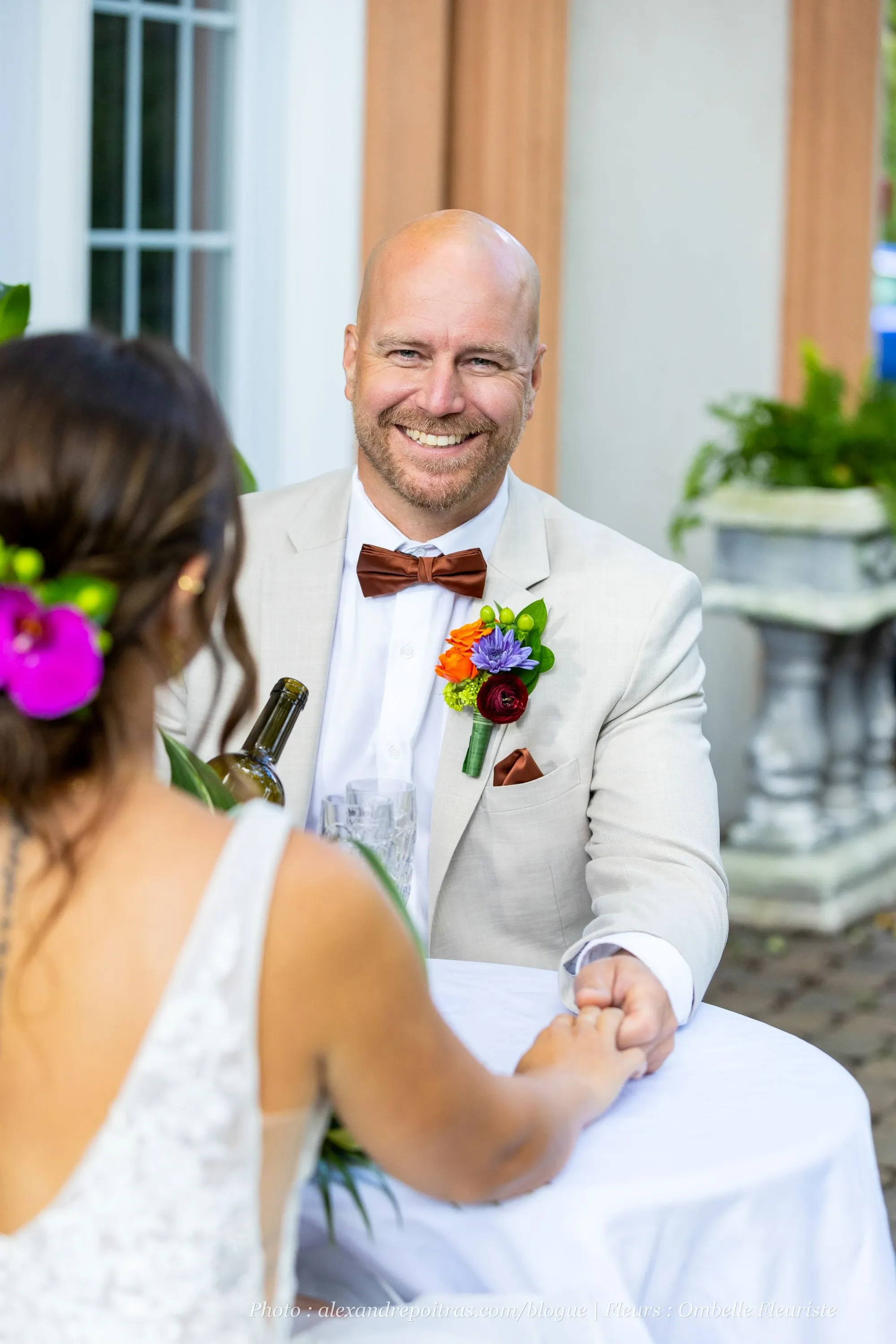 A couple is sitting at an elegantly decorated outdoor wedding reception table. The groom, wearing a light beige suit with a brown bow tie, smiles warmly while holding hands with the bride. He has a vibrant boutonniere featuring a red rose and other colorful flowers, likely arranged by Ombelle Fleuriste, a wedding florist. The bride, with her hair styled in an updo adorned with pink and purple flowers, sits opposite him, wearing a lace wedding gown. The background shows a classic architectural facade with large windows and lush greenery, enhancing the romantic ambiance of the setting.