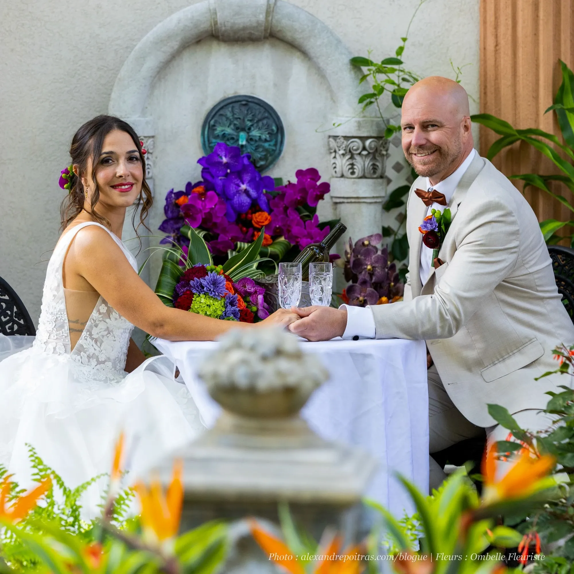A bride and groom are seated at an elegantly decorated table outdoors, with lush greenery and vibrant floral arrangements prominently displayed. The bride wears a beautiful white wedding gown and has a purple flower in her hair, while the groom is dressed in a light-colored suit with a floral boutonniere. The table is adorned with a rich bouquet of purple and red flowers, reflecting the expertise of Ombelle Fleuriste, a vendor specializing in wedding florists for Mariage Elegant in Canada. The setting exudes romance and sophistication, creating a perfect backdrop for a wedding celebration.
