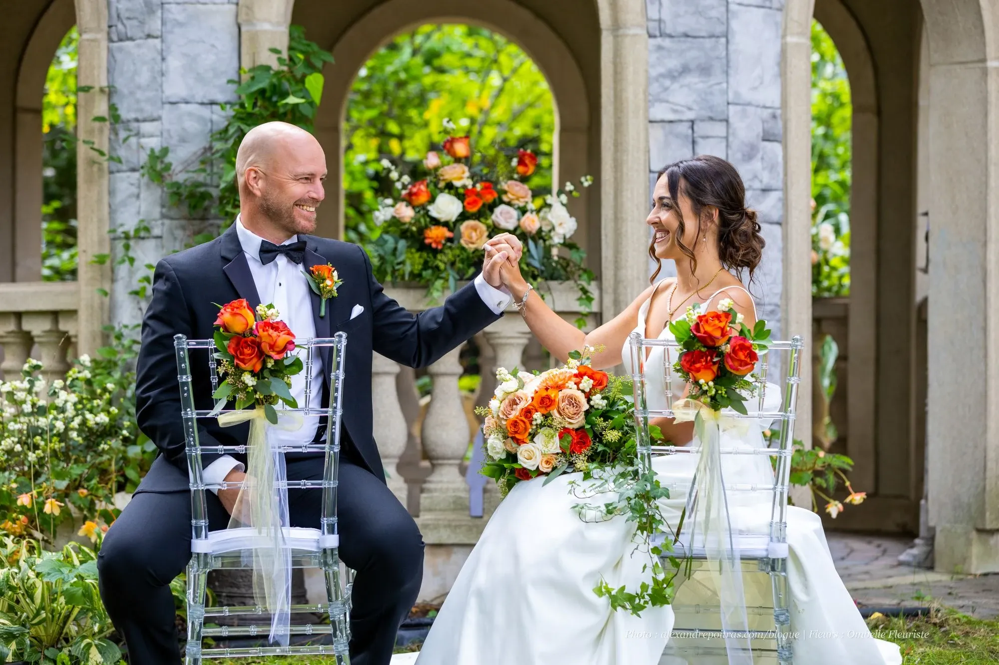A joyful couple sits on clear chairs at an elegantly decorated stone terrace, surrounded by lush greenery and colorful floral arrangements. The groom is wearing a black tuxedo adorned with a vibrant orange boutonniere, while the bride is dressed in a stunning white gown. Both chairs are beautifully decorated with bouquets of orange and white flowers. An exquisite floral arrangement by Ombelle Fleuriste graces the background, enhancing the romantic ambiance. This image captures the essence of Ombelle Fleuriste's expertise as a wedding florist, showcasing their talent in creating breathtaking wedding decor for Mariage Elegant in Canada.