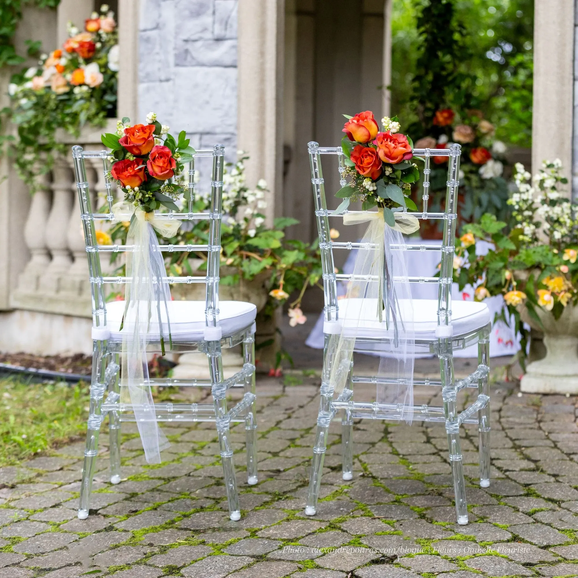 This image features an elegant outdoor wedding setup showcasing the floral artistry of Ombelle Fleuriste, a prominent vendor in the Wedding Florists category. It depicts two transparent chairs adorned with vibrant orange and red rose arrangements, accented by delicate green foliage. These beautiful bouquets are tied to the chairs with soft, flowing white ribbons, adding a touch of sophistication. The backdrop includes a stone structure adorned with lush greenery and additional flower arrangements, creating a romantic and inviting atmosphere perfect for a wedding celebration.