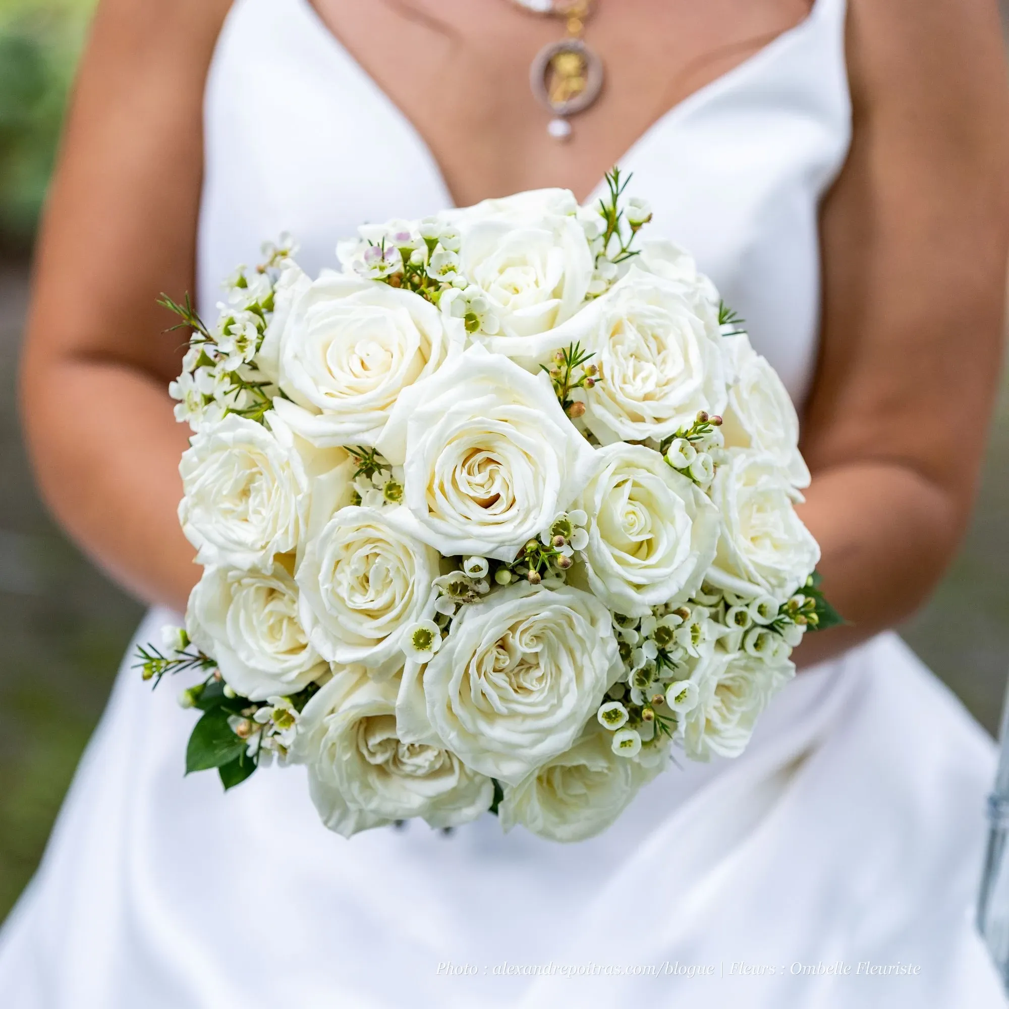 The image features a bride holding a stunning bouquet by Ombelle Fleuriste, a renowned wedding florist, perfect for elegant weddings. The bouquet consists of lush white roses intertwined with delicate greenery and small white flowers, creating a classic and sophisticated look ideal for wedding ceremonies. The bride's gown is white, complementing the floral arrangement, and she wears a pendant necklace, all capturing the essence of timeless bridal elegance.