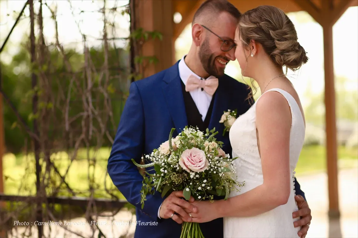 In this image, a newlywed couple stands closely together in an outdoor setting, showcasing a romantic moment. The groom, wearing a blue suit and pink bow tie, lovingly gazes at the bride, who is dressed in a white lace wedding gown. She holds a bouquet designed by Ombelle Fleuriste, a vendor specializing in wedding florists, featuring soft pink roses, white baby's breath, and green foliage. The wooden pergola and greenery in the background add to the serene and elegant wedding atmosphere, suitable for Mariage Elegant's planning and vendor listing website in Canada.