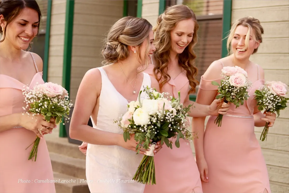 The image features a joyful bridal party walking together, with the bride in a white lace wedding dress holding a bouquet filled with white roses and greenery, designed by Ombelle Fleuriste. Surrounding her are three bridesmaids in matching blush pink dresses, each holding smaller bouquets of pink roses and baby's breath. The setting appears to be a charming, rustic venue with wooden siding, perfectly capturing a moment of celebration and elegance. Ombelle Fleuriste showcases their expertise in wedding floristry through these beautifully arranged floral bouquets.