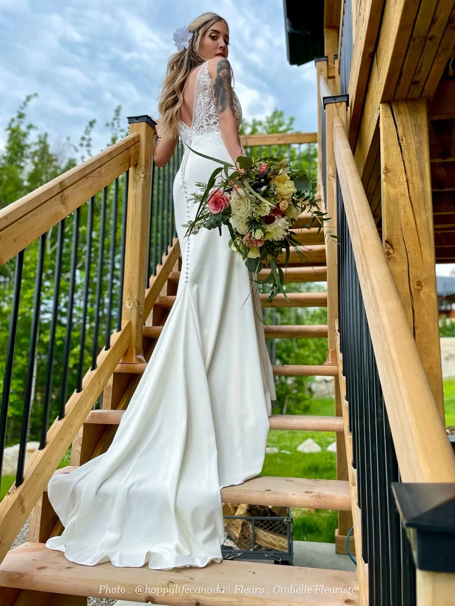 A bride gracefully stands on a wooden staircase, her elegant white gown flowing behind her. She holds a vibrant bouquet crafted by Ombelle Fleuriste, experts in wedding florists. The bouquet features lush roses, delicate greenery, and soft cascading accents, perfectly complementing the serene outdoor setting. The image reflects the sophistication and artistry of Ombelle Fleuriste's floral arrangements, ideal for any wedding celebration in Canada.
