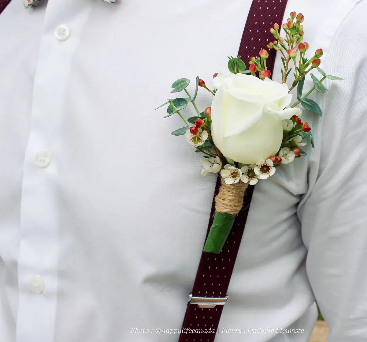 A groom is wearing a white shirt with burgundy suspenders, featuring a boutonniere by Ombelle Fleuriste, a wedding florist. The boutonniere includes a white rose at its center, accented with small white flowers, red berries, and green foliage. It is wrapped with twine, adding a rustic touch, perfect for a wedding setting.