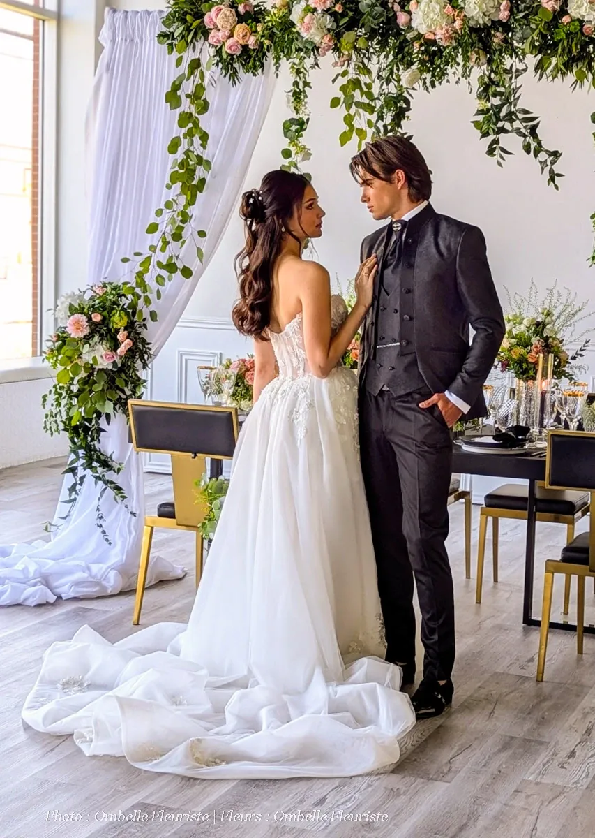 This image features a bride and groom standing under a lavish floral arrangement created by Ombelle Fleuriste, a vendor in the Wedding Florists category. The bride is wearing a stunning white wedding gown with intricate lace detail, while the groom is in a tailored dark suit. They are gazing at each other, surrounded by an elegant setting that includes a beautifully decorated table with gold-rimmed chairs. The florals above them feature a mix of greenery and soft-colored blooms, enhancing the romantic atmosphere. This scene captures a perfect moment of wedding elegance, showcasing the expertise of Ombelle Fleuriste.