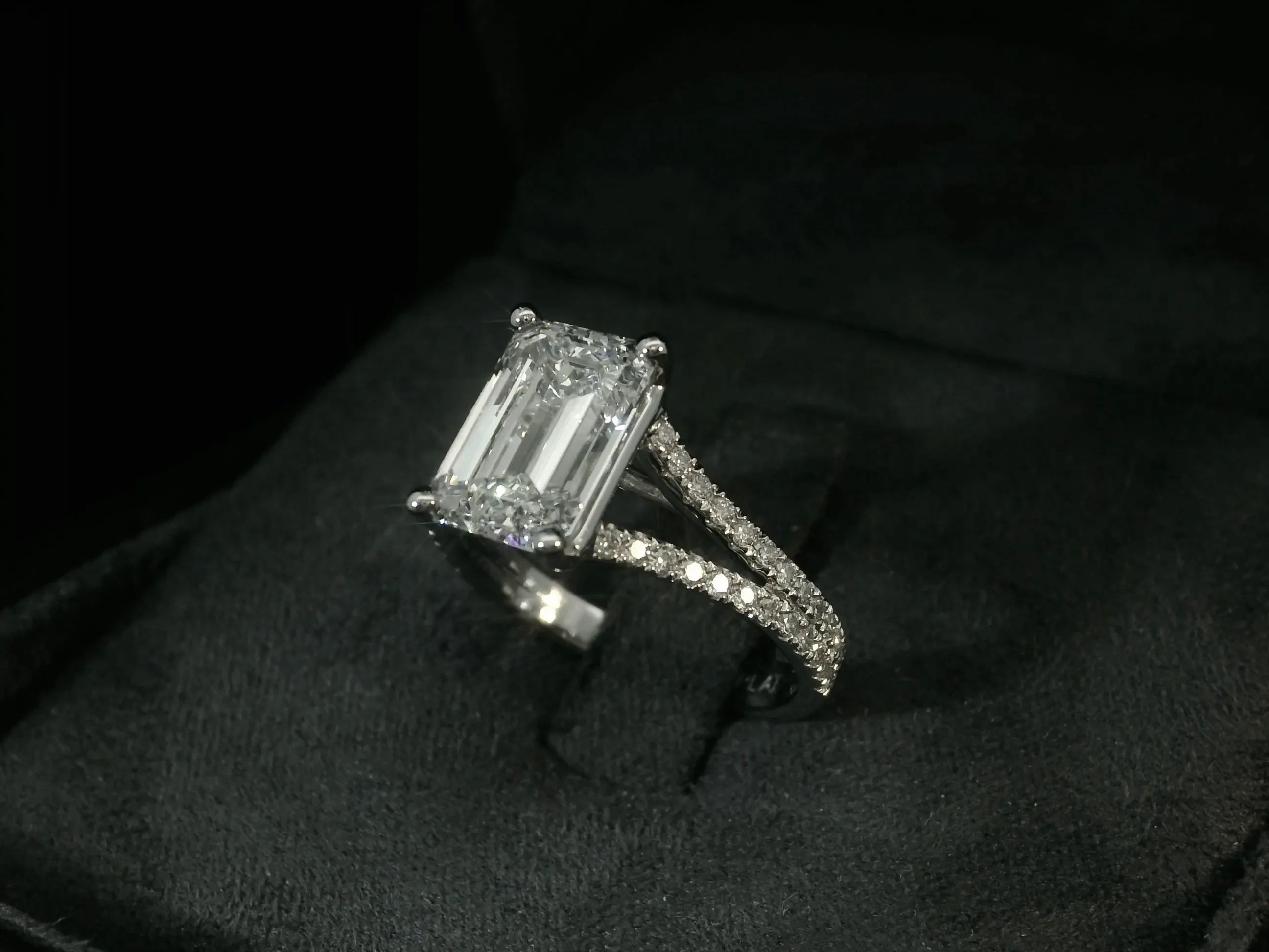 This image displays a stunning engagement ring from Angelica Jewellers, featuring in the wedding jewelry category on Mariage Elegant, Canada. The ring showcases a large, emerald-cut diamond set prominently on a sleek, silver band. The band is adorned with smaller, round-cut diamonds that add an extra layer of sparkle and sophistication. The ring is elegantly positioned on a plush, black velvet box, highlighting its exquisite craftsmanship and attention to detail, making it an ideal choice for those seeking elegant wedding jewelry.