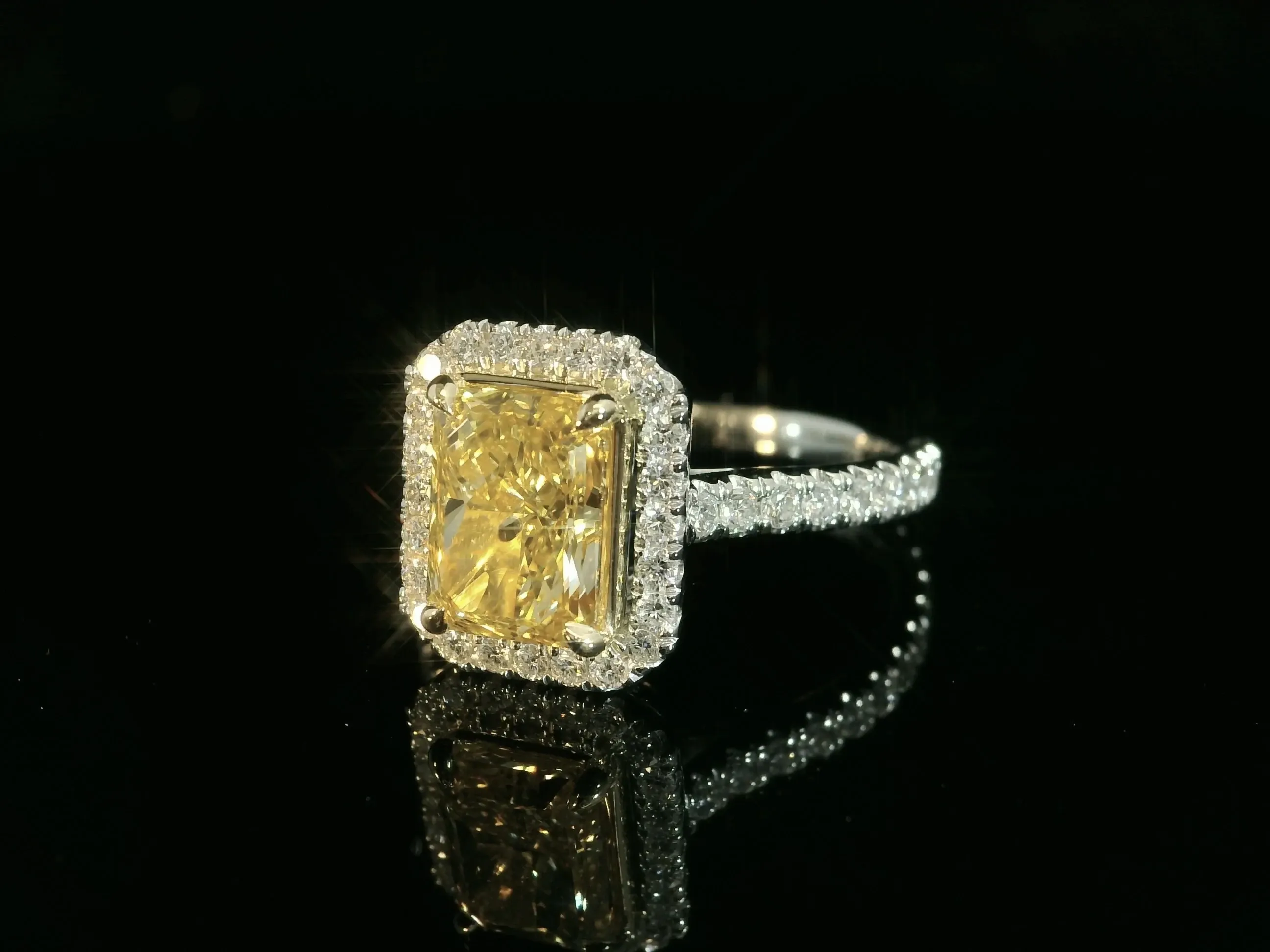 This image features a stunning engagement ring designed by Angelica Jewellers, showcased under the wedding jewelry category. The ring prominently displays a large, radiant-cut yellow diamond at its center, encircled by a halo of exquisite white diamonds. The intricate band also glitters with a series of smaller white diamonds, enhancing the ring's overall brilliance. The elegant and luxurious design epitomizes the exquisite craftsmanship suitable for a sophisticated wedding setting, reflecting the high-quality offerings available through Mariage Elegant in Canada.