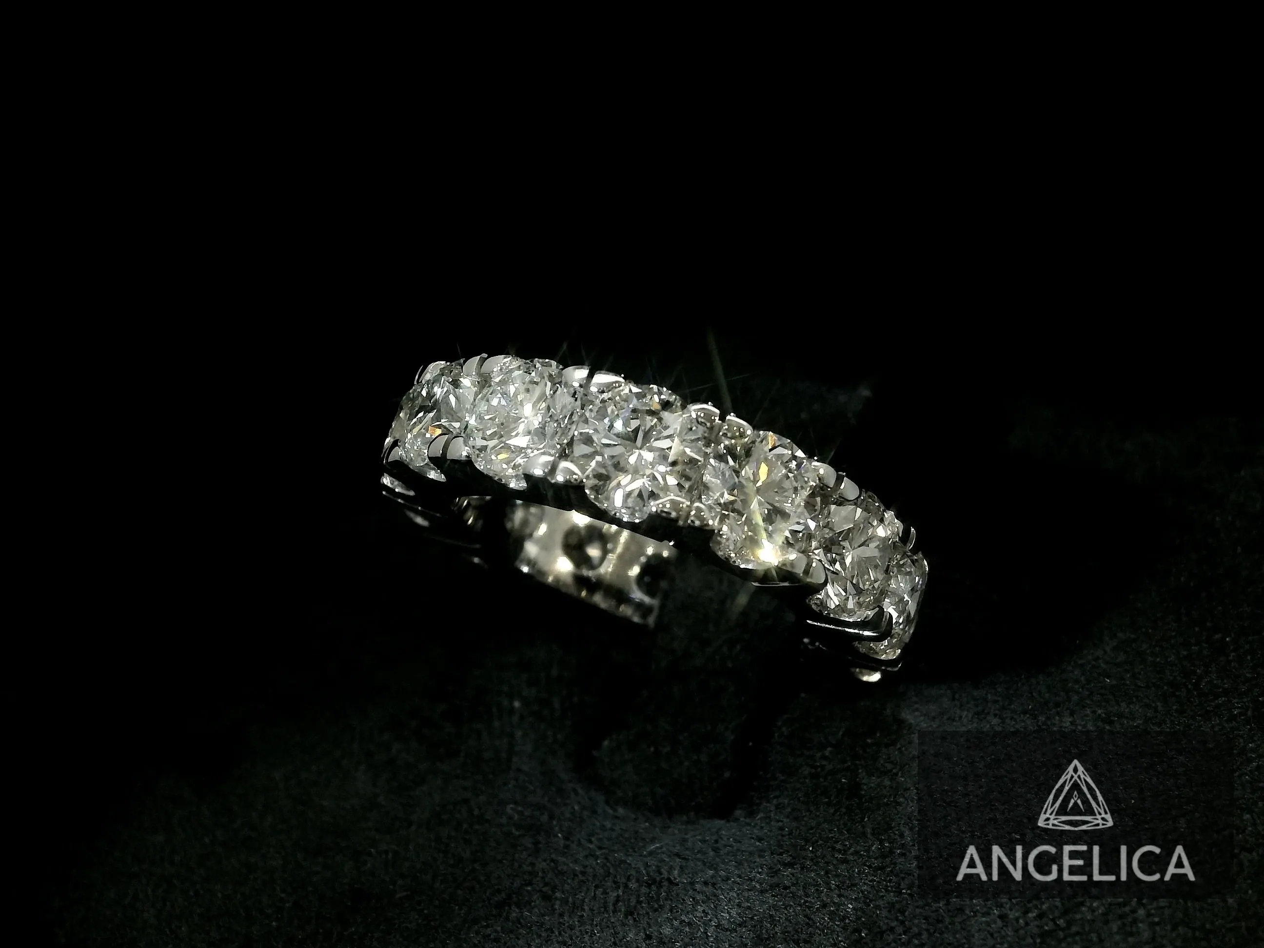 This image features a stunning wedding jewelry piece by Angelica Jewellers, displaying a beautifully crafted silver band adorned with a series of sparkling, round-cut diamonds. The elegant design catches the light, emphasizing the brilliance and clarity of each stone. Set against a dark background, the focus is entirely on the exquisite craftsmanship and luxurious appeal of the ring, suitable for a sophisticated wedding setting. The Angelica Jewellers logo is subtly visible in the bottom right corner, reinforcing the brand's association with high-quality wedding jewelry.