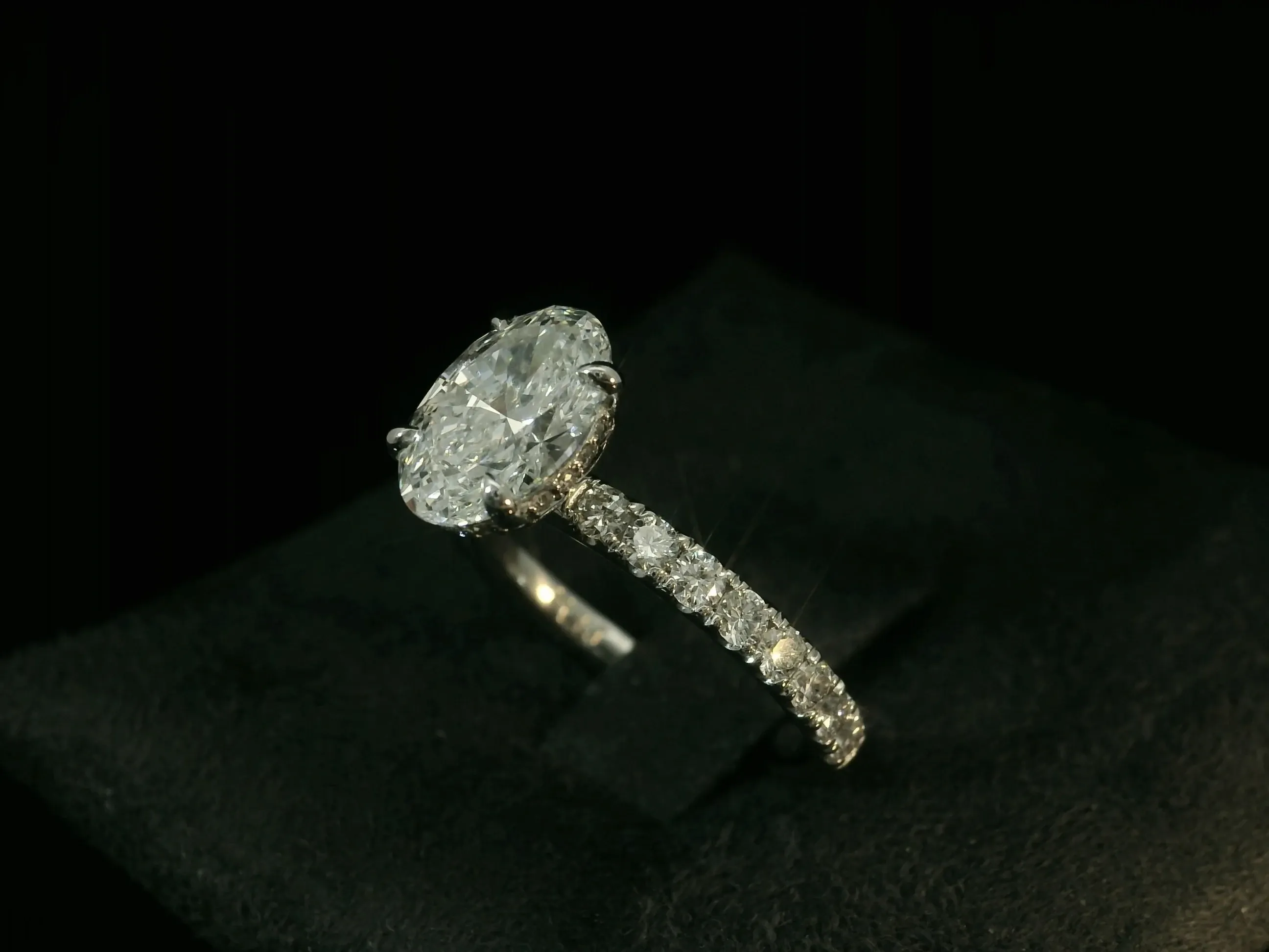 A stunning diamond engagement ring by Angelica Jewellers, featured in the wedding jewelry category. The ring showcases a large, oval-cut diamond set in a prong setting, complemented by a band encrusted with smaller sparkling diamonds. The elegant jewelry piece is displayed on a dark, textured background, highlighting its brilliance and craftsmanship, perfect for a timeless wedding proposal.