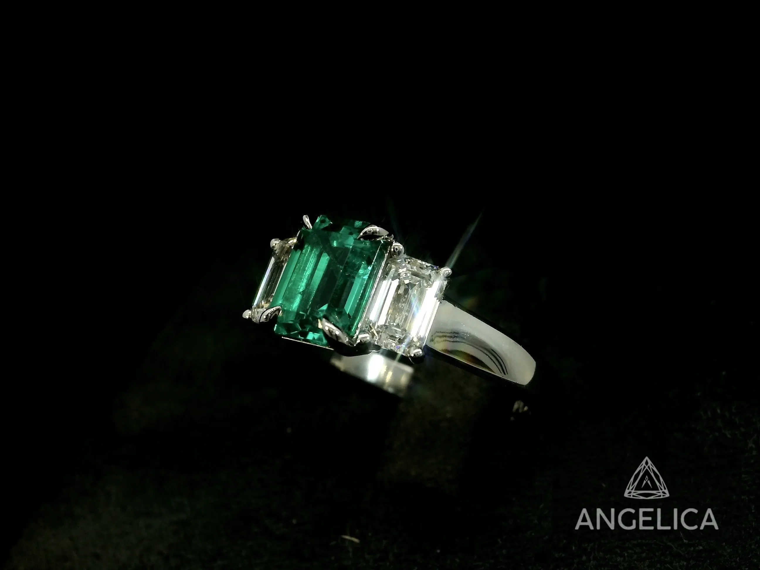 This image showcases a stunning emerald engagement ring from Angelica Jewellers, a vendor in the Wedding Jewelry category in Canada. The ring features a vibrant green emerald cut gemstone set in the center, flanked by two sparkling rectangular diamonds on either side. The simple, elegant band is made of polished white gold, highlighting the brilliance of the stones. The jeweller's branding, 'ANGELICA,' is subtly displayed in the corner, adding a touch of elegance and authenticity to the piece. This exquisite ring is perfect for those seeking a luxurious and unique statement piece for their wedding.