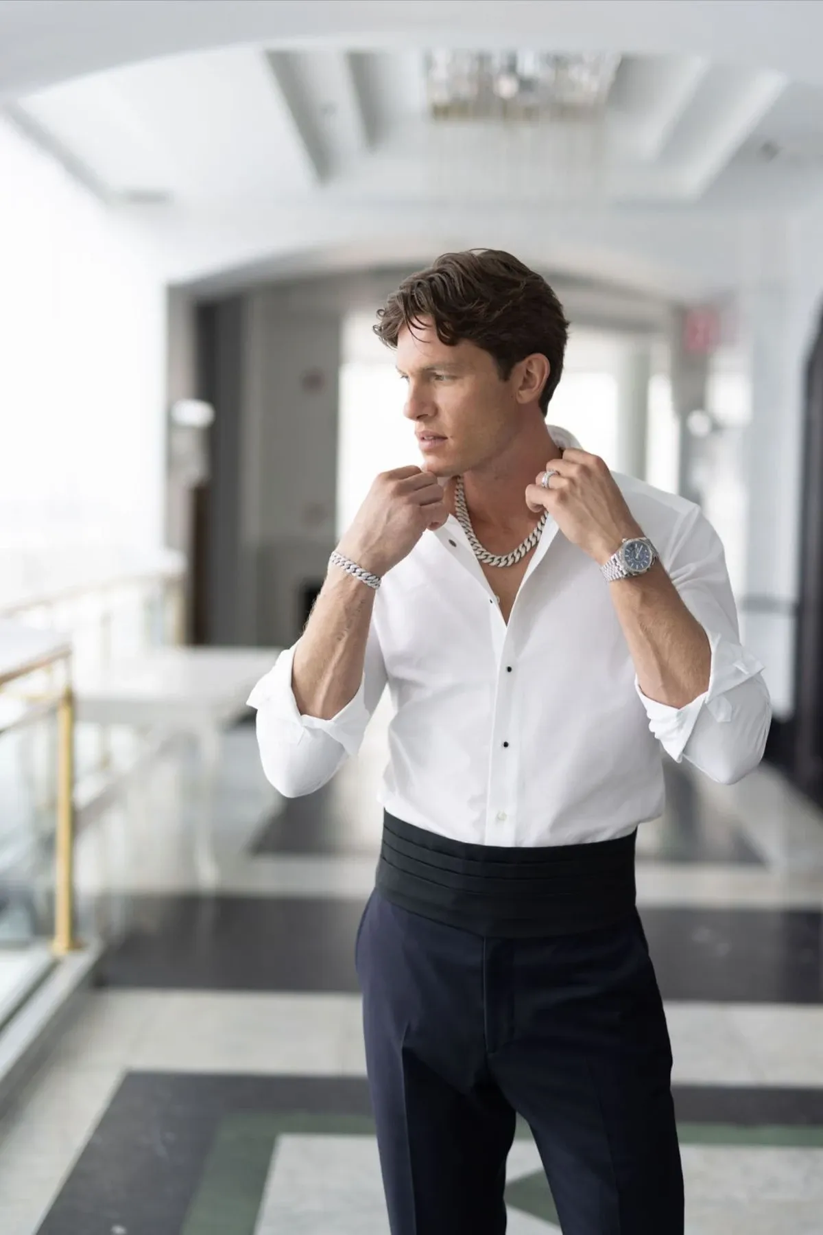 A groom standing in a bright, elegant hallway is adjusting his cufflinks while wearing a classic white dress shirt and dark formal trousers. The groom is adorned with a sophisticated silver chain necklace and a sleek wristwatch, both designed by Angelica Jewellers, a featured vendor in the Wedding Jewelry category at Mariage Elegant, Canada. The setting exudes luxury and refinement, reflecting a sophisticated wedding atmosphere.