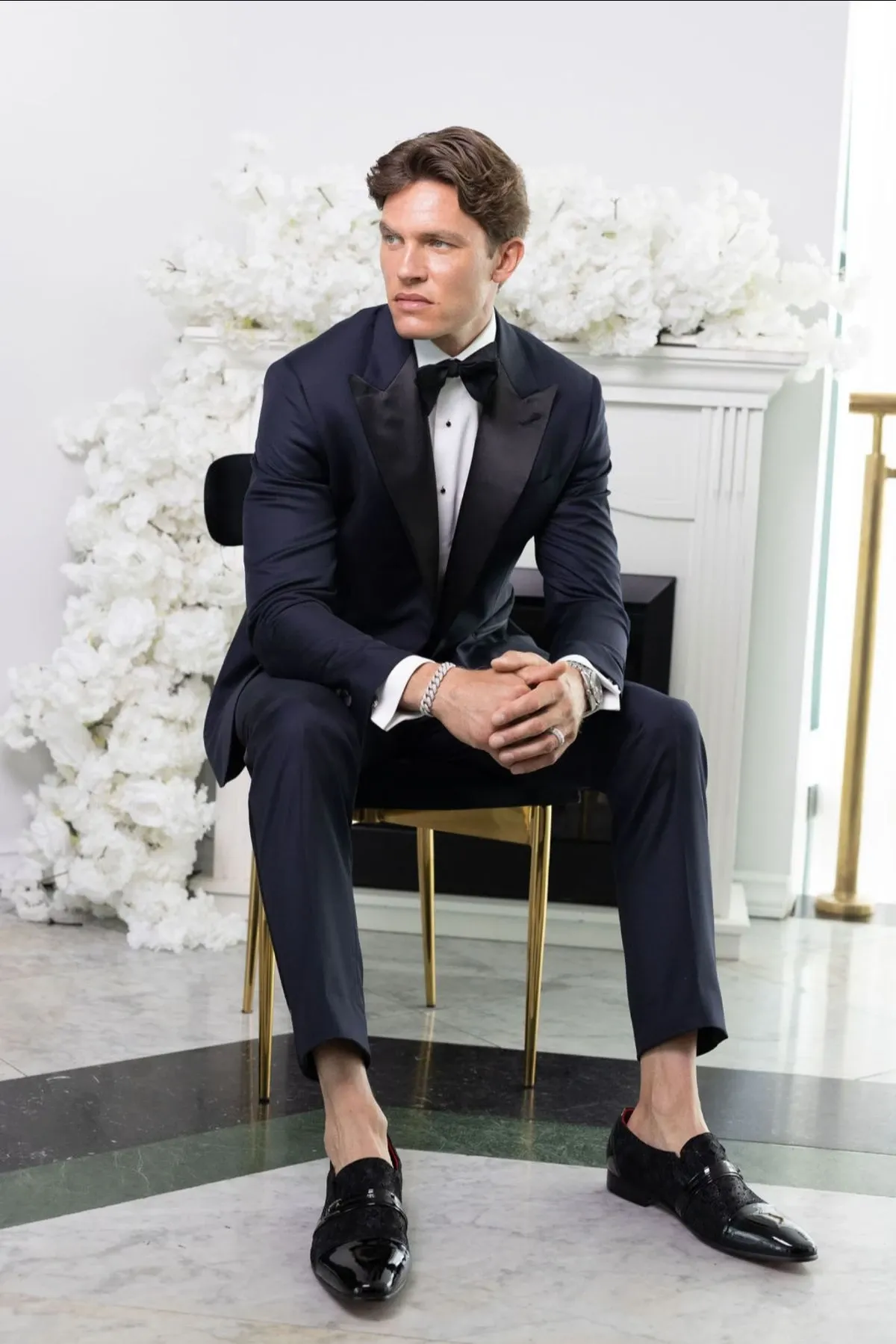 A groom is seated in an elegant setting, wearing a sophisticated black tuxedo with a bow tie. The focal point is the jewelry from Angelica Jewellers, highlighting a refined bracelet that complements his attire. Behind him, a white floral arrangement adds a touch of wedding elegance, enhancing the luxurious atmosphere. This image showcases wedding jewelry in the context of Mariage Elegant's vendor listing for wedding planning in Canada.