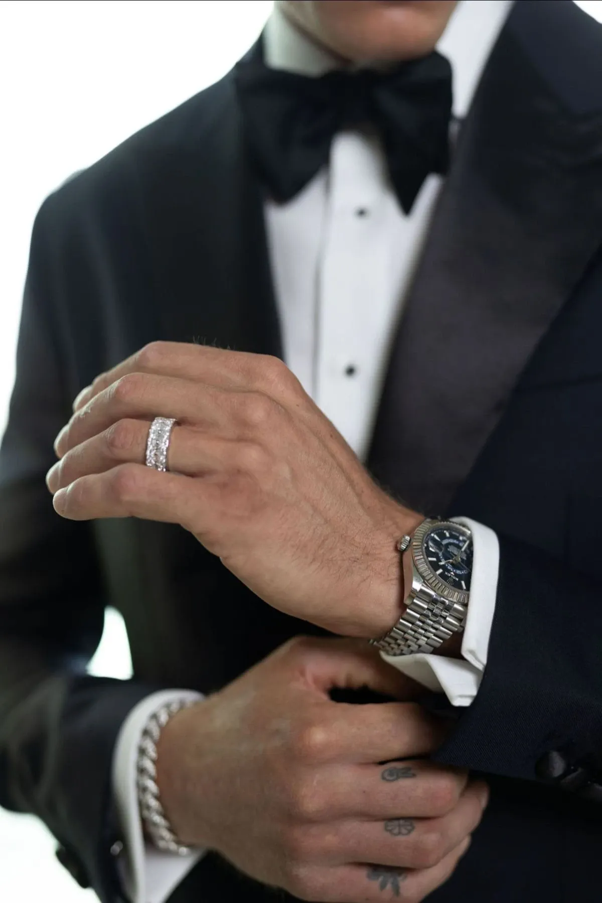 A groom elegantly dressed in a tailored black tuxedo is adjusting his watch, showcasing a finely detailed wedding band and a matching bracelet. The jewelry, provided by Angelica Jewellers, is category-defining for wedding jewelry with its sophisticated and timeless design, ideal for a stylish wedding.
