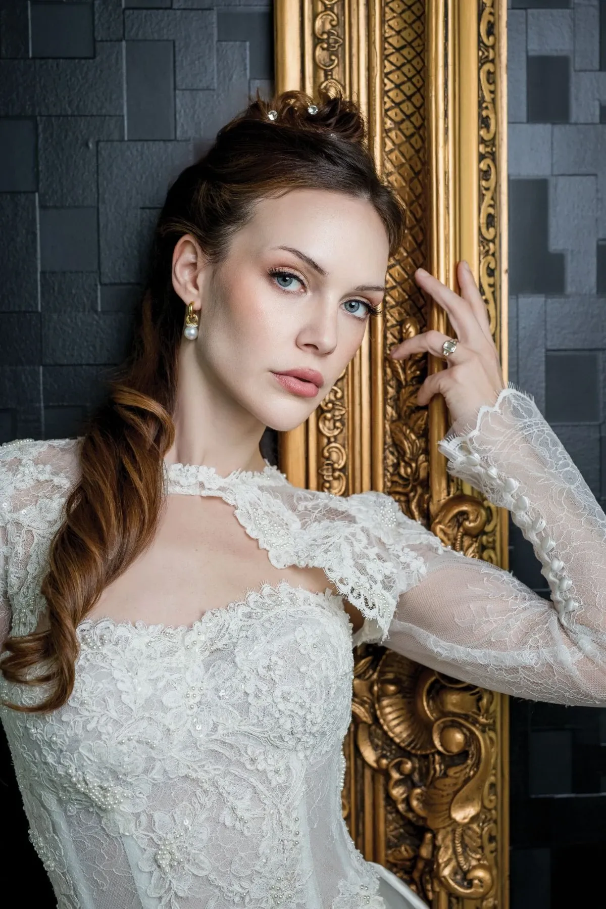 A bride is elegantly posed against a luxurious gold ornate backdrop, showcasing exquisite wedding jewelry by Angelica Jewellers. She is wearing a delicate lace bridal gown, highlighting her sophisticated style. Her hair is styled in soft waves adorned with small, subtle accessories. The focus is on her sparkling earrings and an engagement ring, which are perfect examples of Angelica Jewellers' wedding jewelry offerings, ideal for adding a touch of glamour to any wedding ensemble.